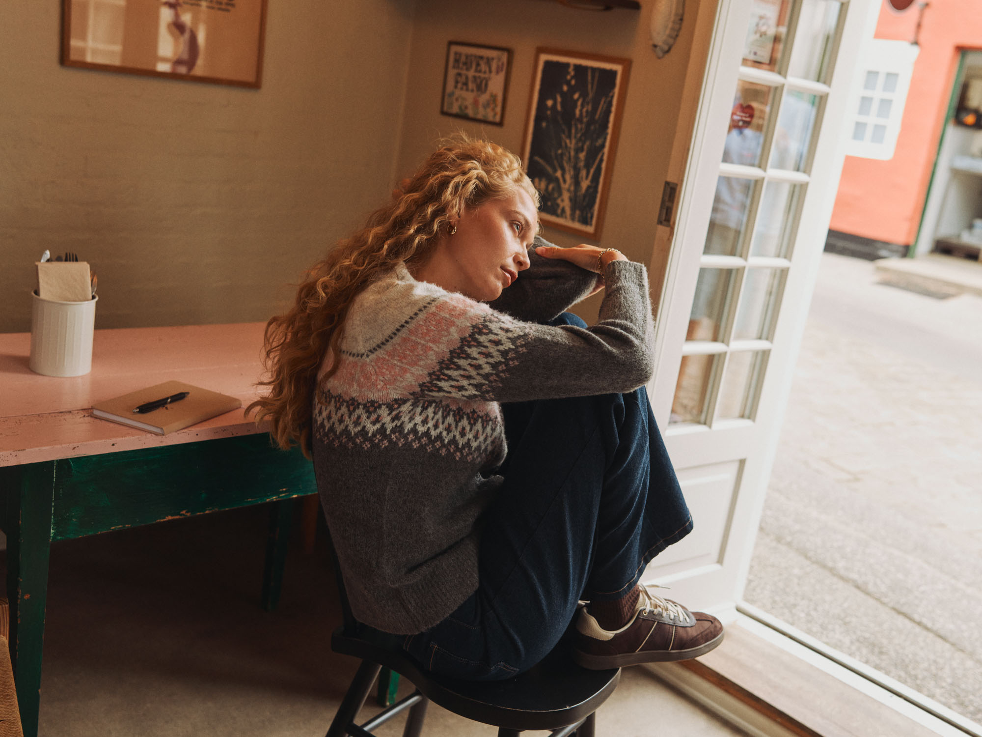 Curly-haired woman in fair isle sweater sits, knees hugged, looking out an open door.