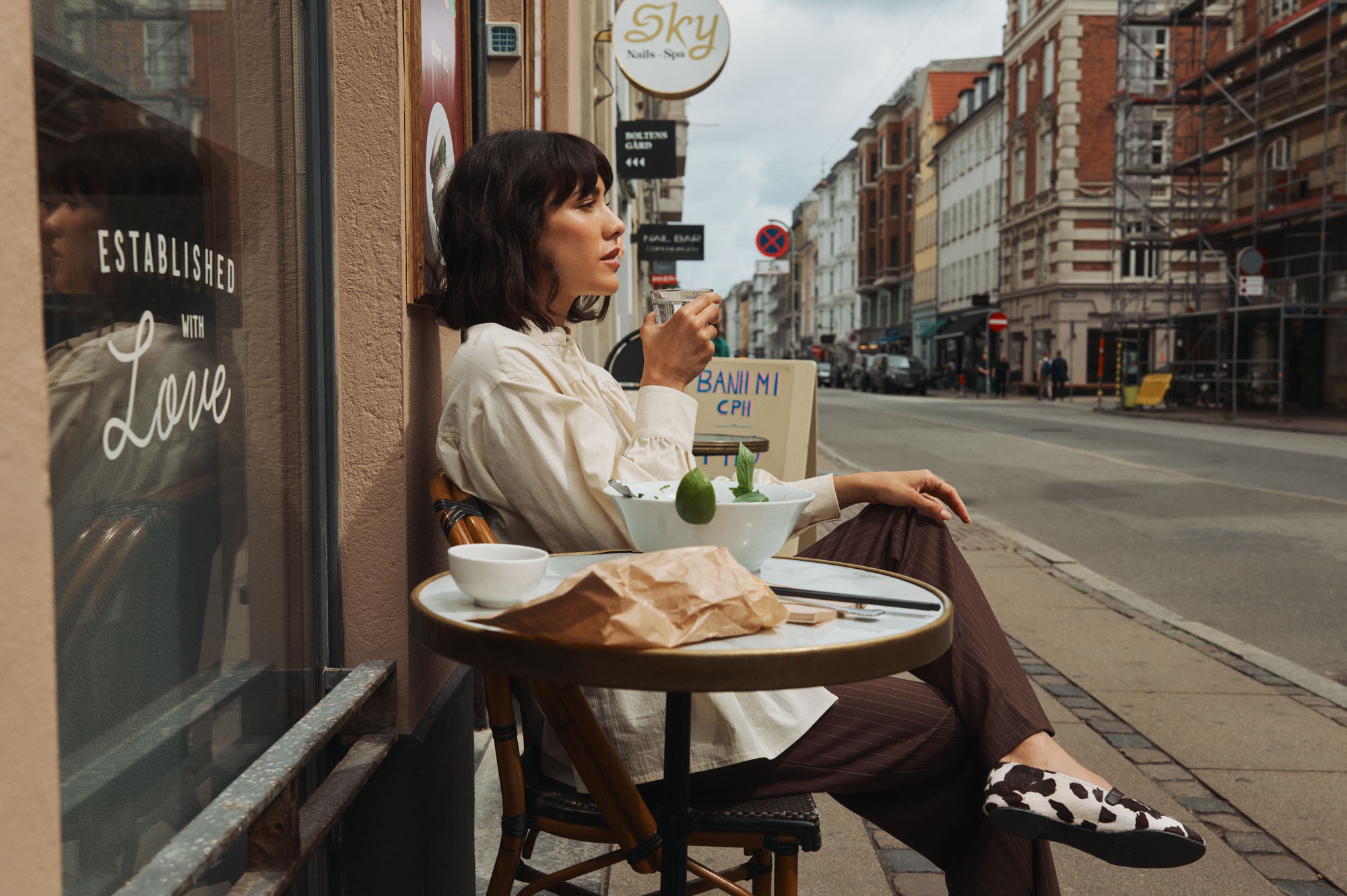 Woman in cream shirt sips drink at outdoor cafe table with food, city street background.