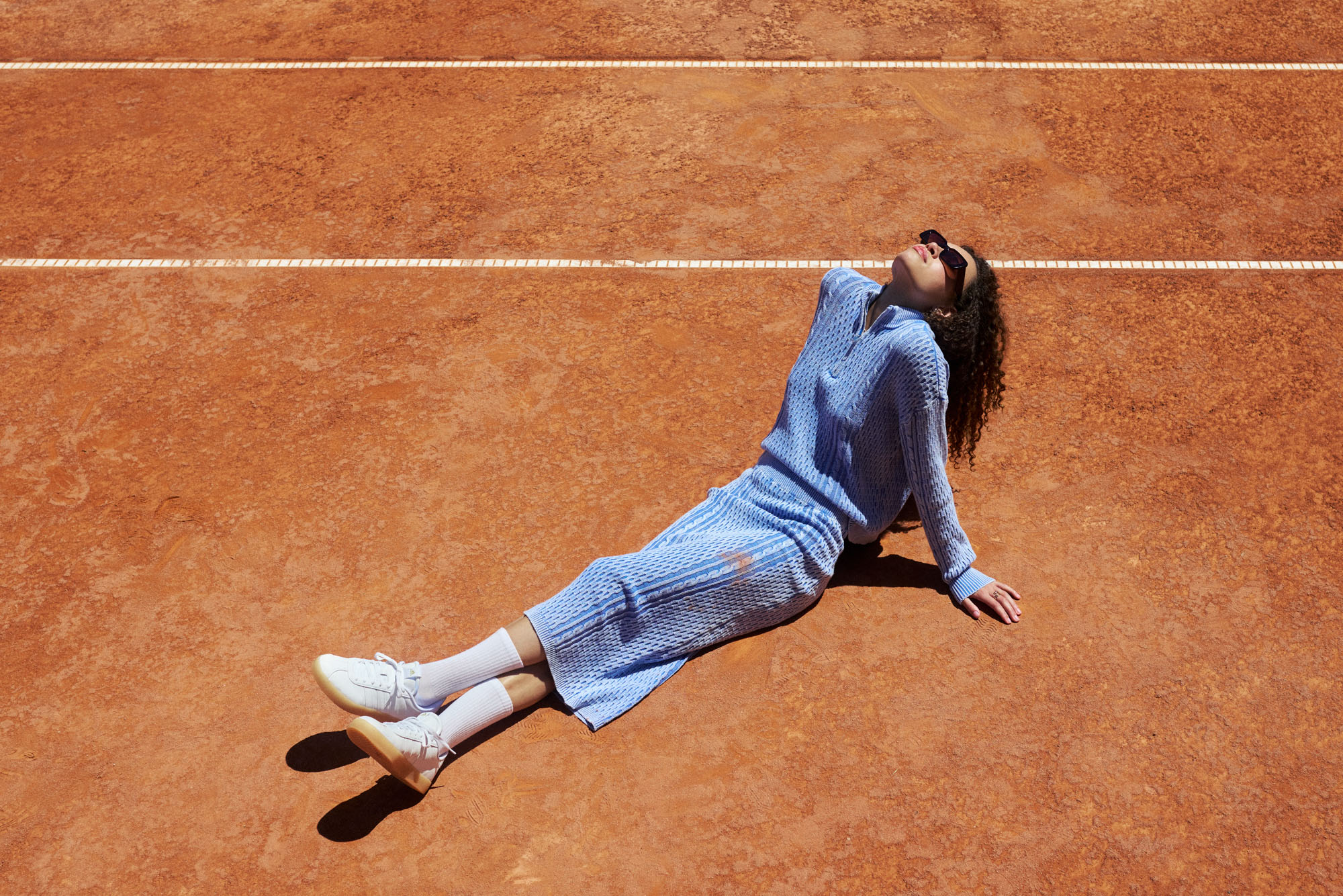 Person in blue knitwear and white sneakers relaxing on a sunny clay tennis court