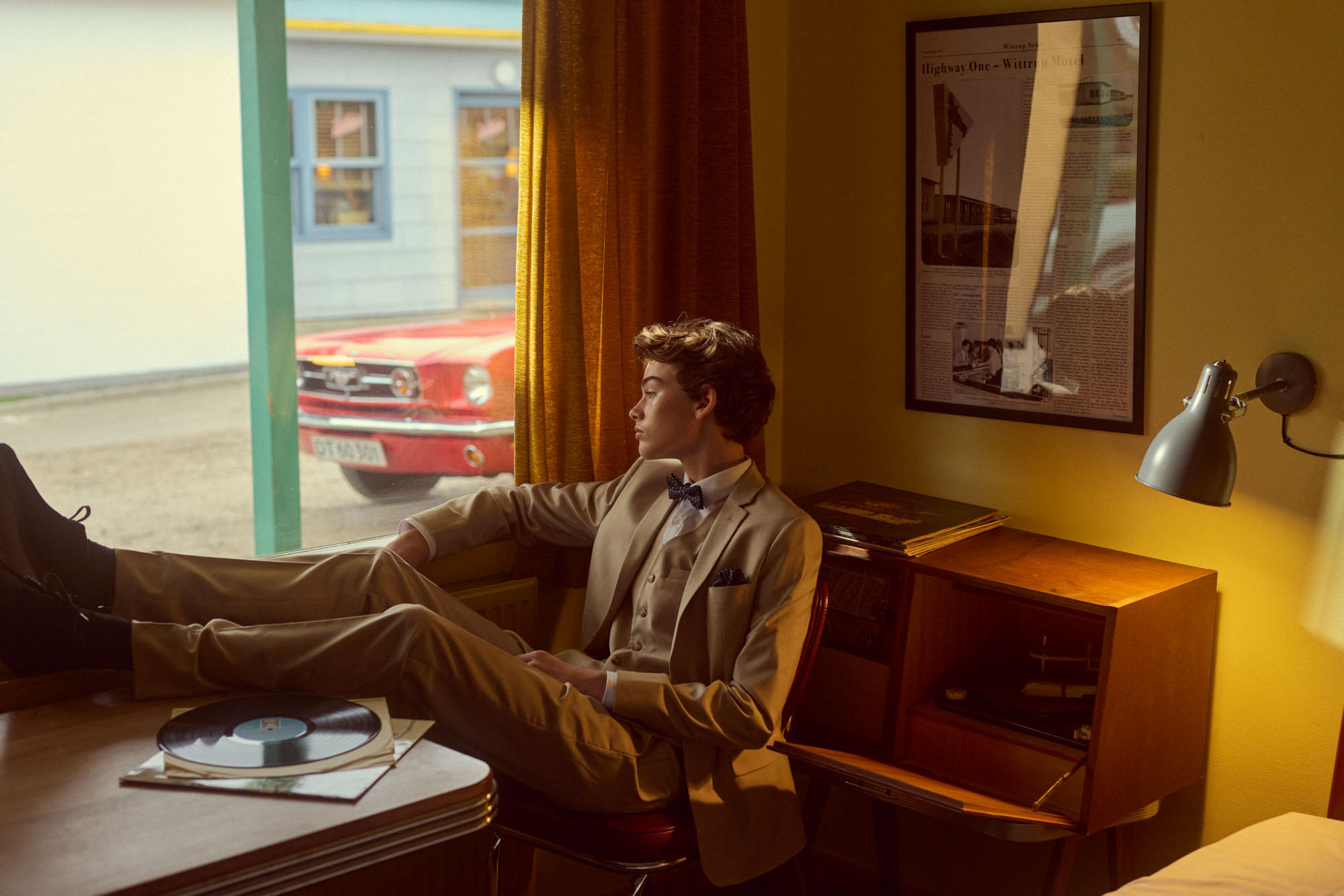 Stylish young man in tan suit sits in retro room, gazing at classic red car.