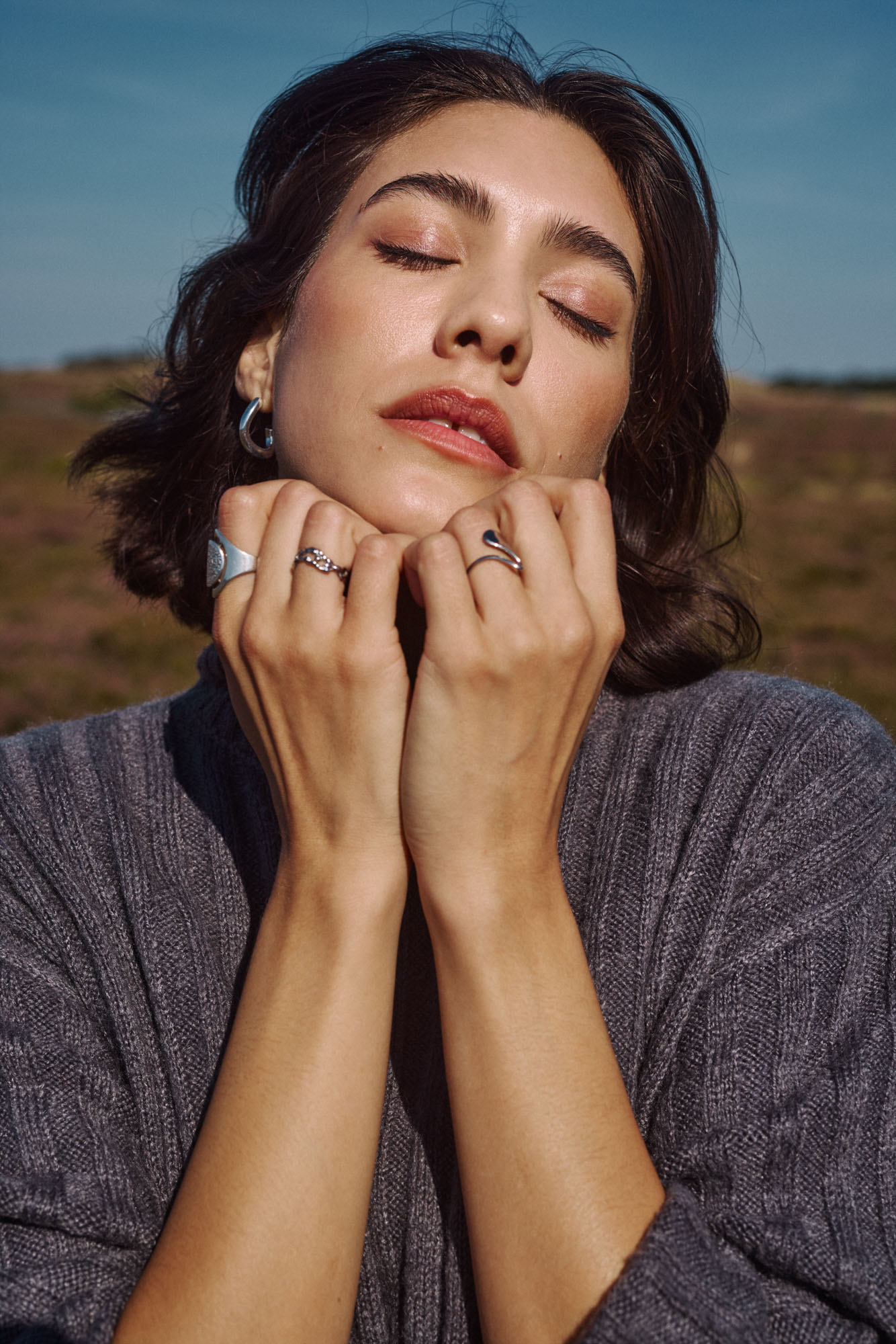 Close-up of a serene woman with closed eyes, wearing rings and a grey sweater outdoors.