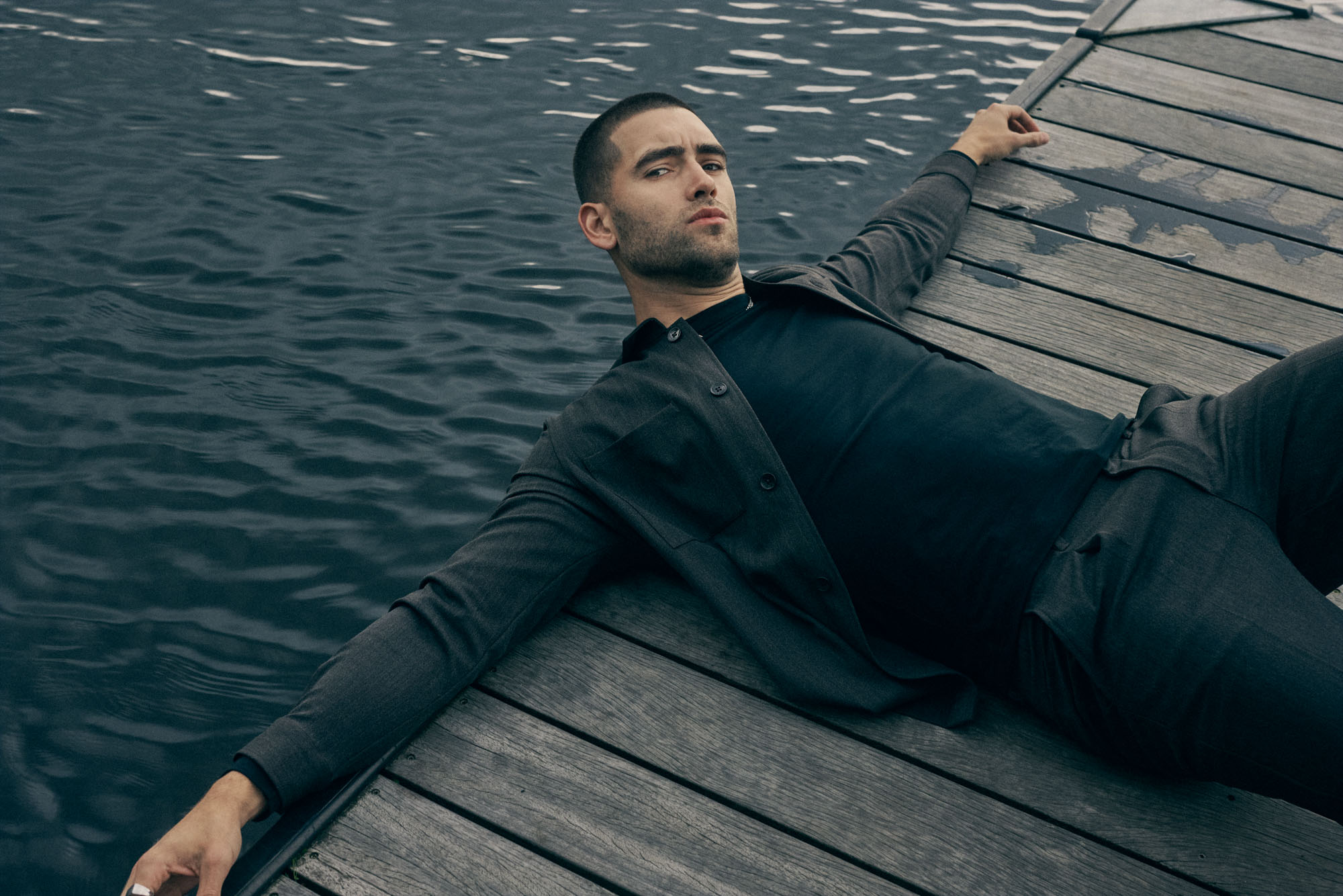 Man in dark clothing lies on a wooden dock by dark water, gazing at viewer.