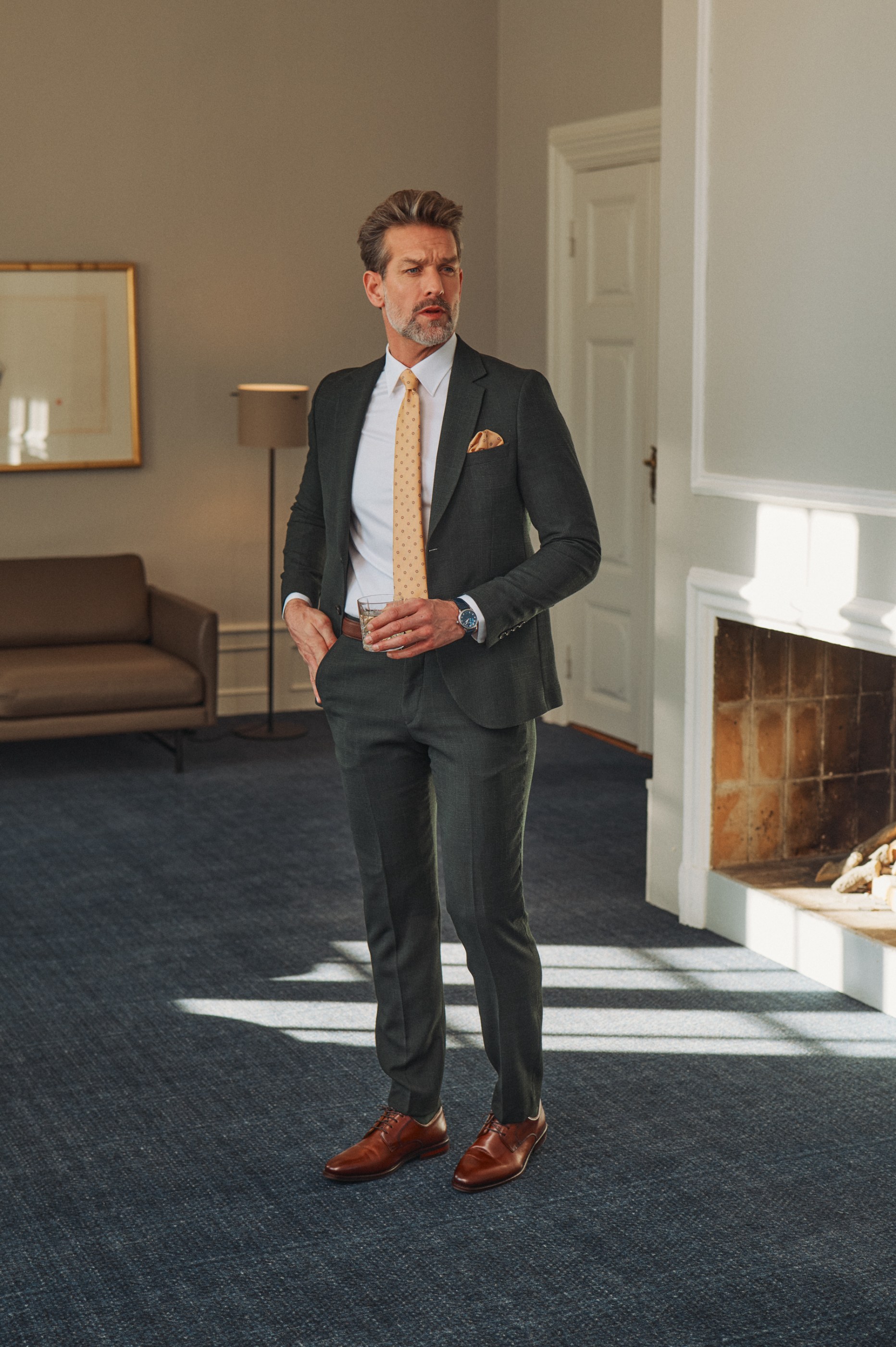 Stylish man in a dark green suit and yellow tie holding a drink in a sunlit room.