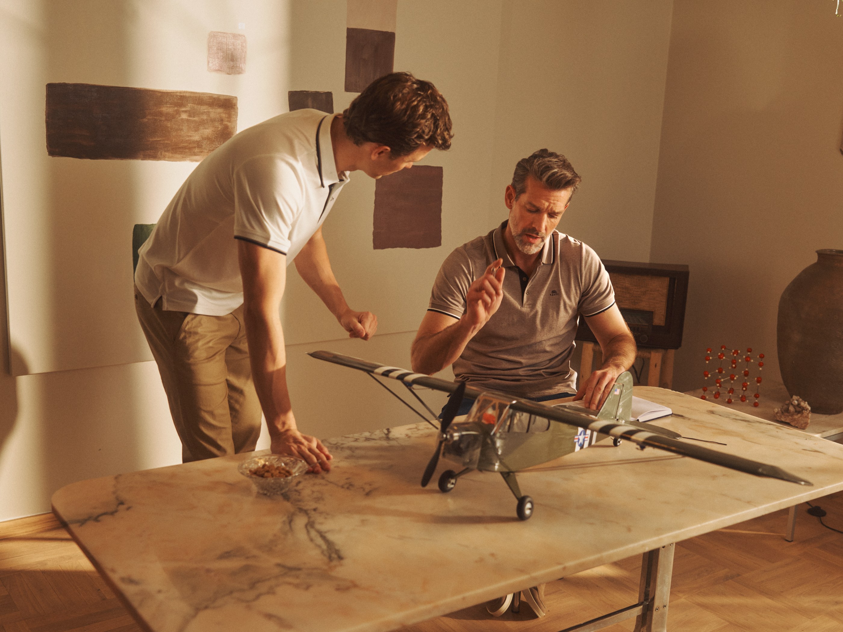 Two men in polo shirts intently working on a model airplane at a marble table.