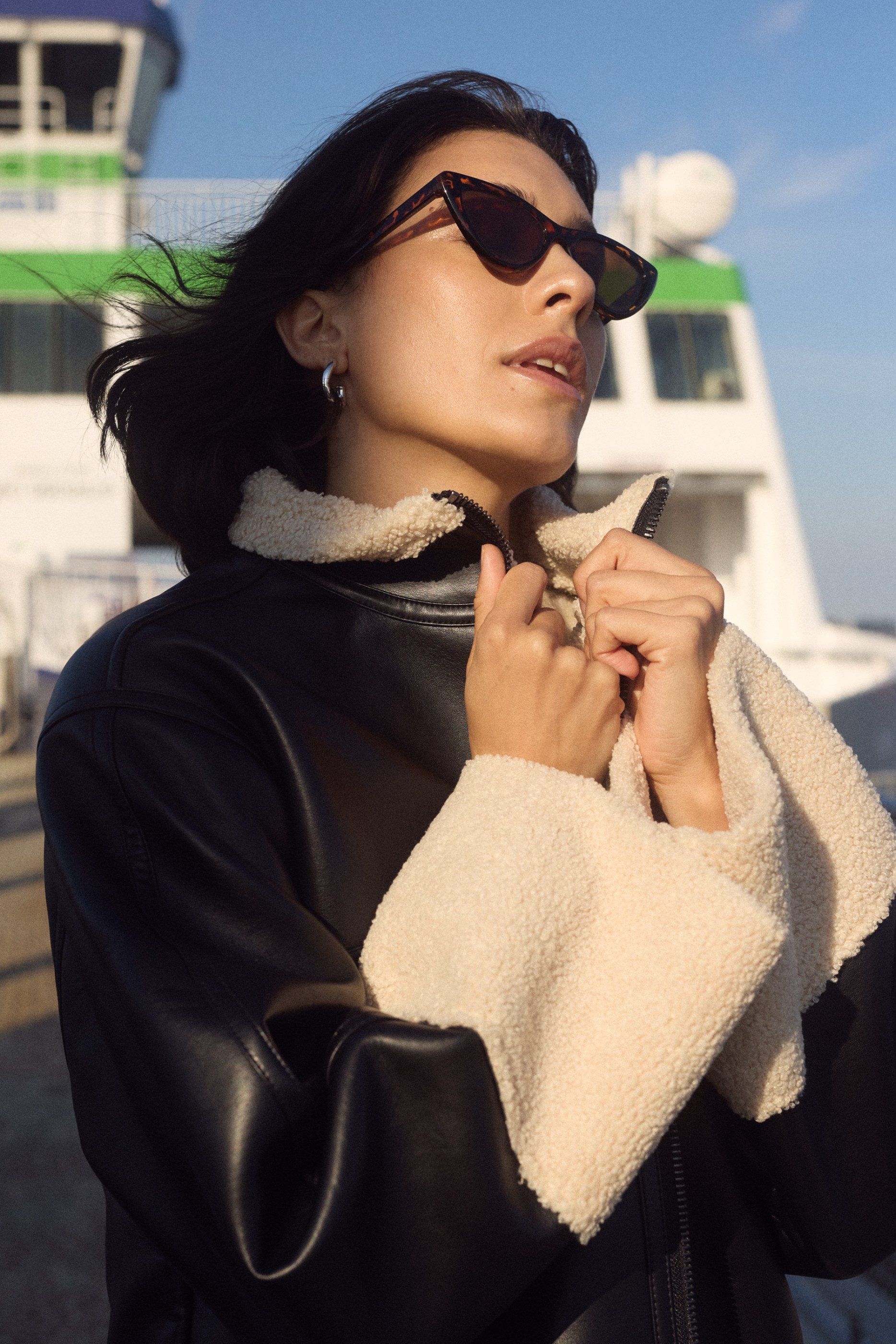 Woman adjusting black leather and cream sherpa jacket, wearing sunglasses, on a boat.