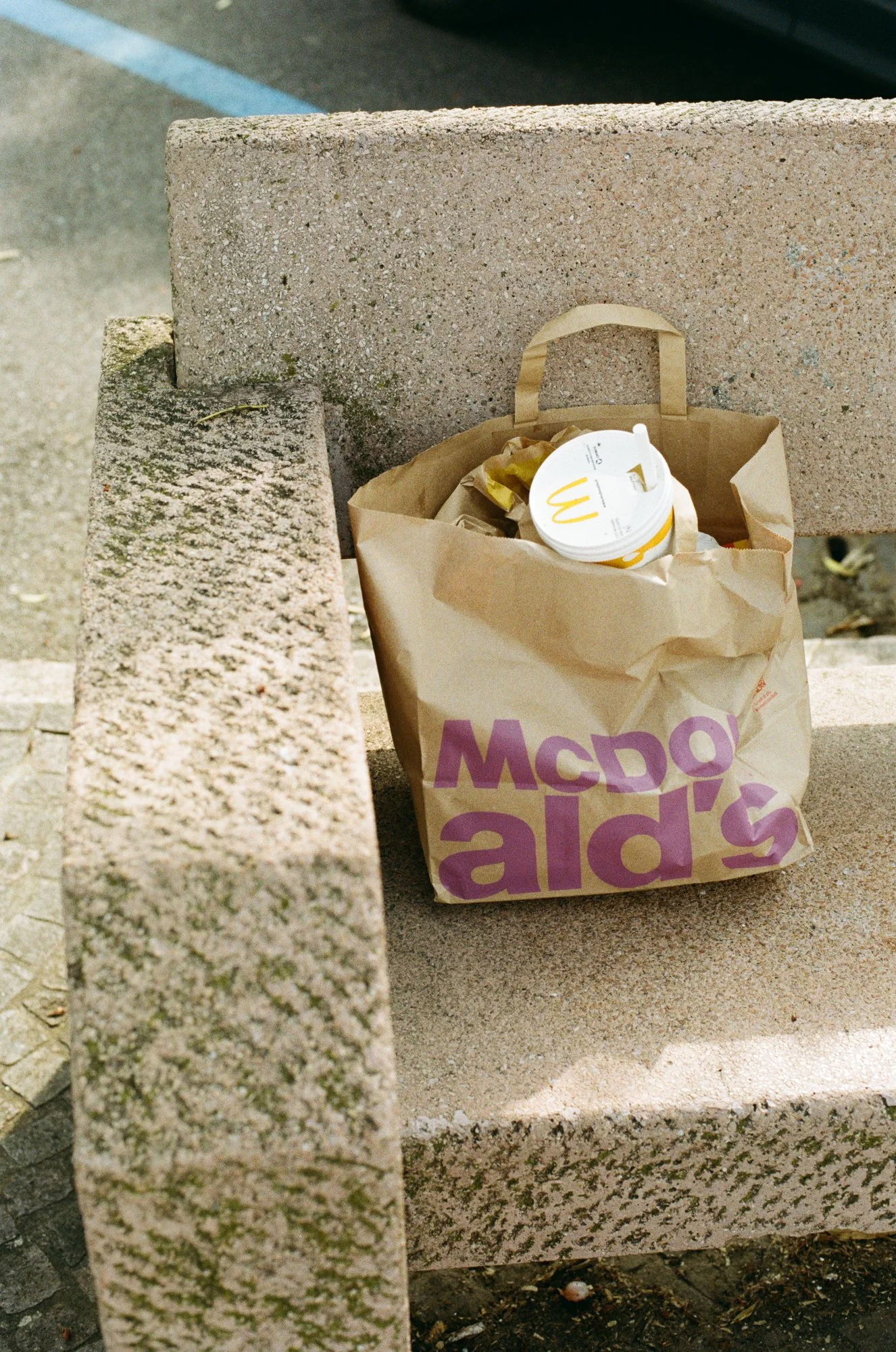 A McDonald's take-out bag with a drink cup rests on a sunny concrete bench.
