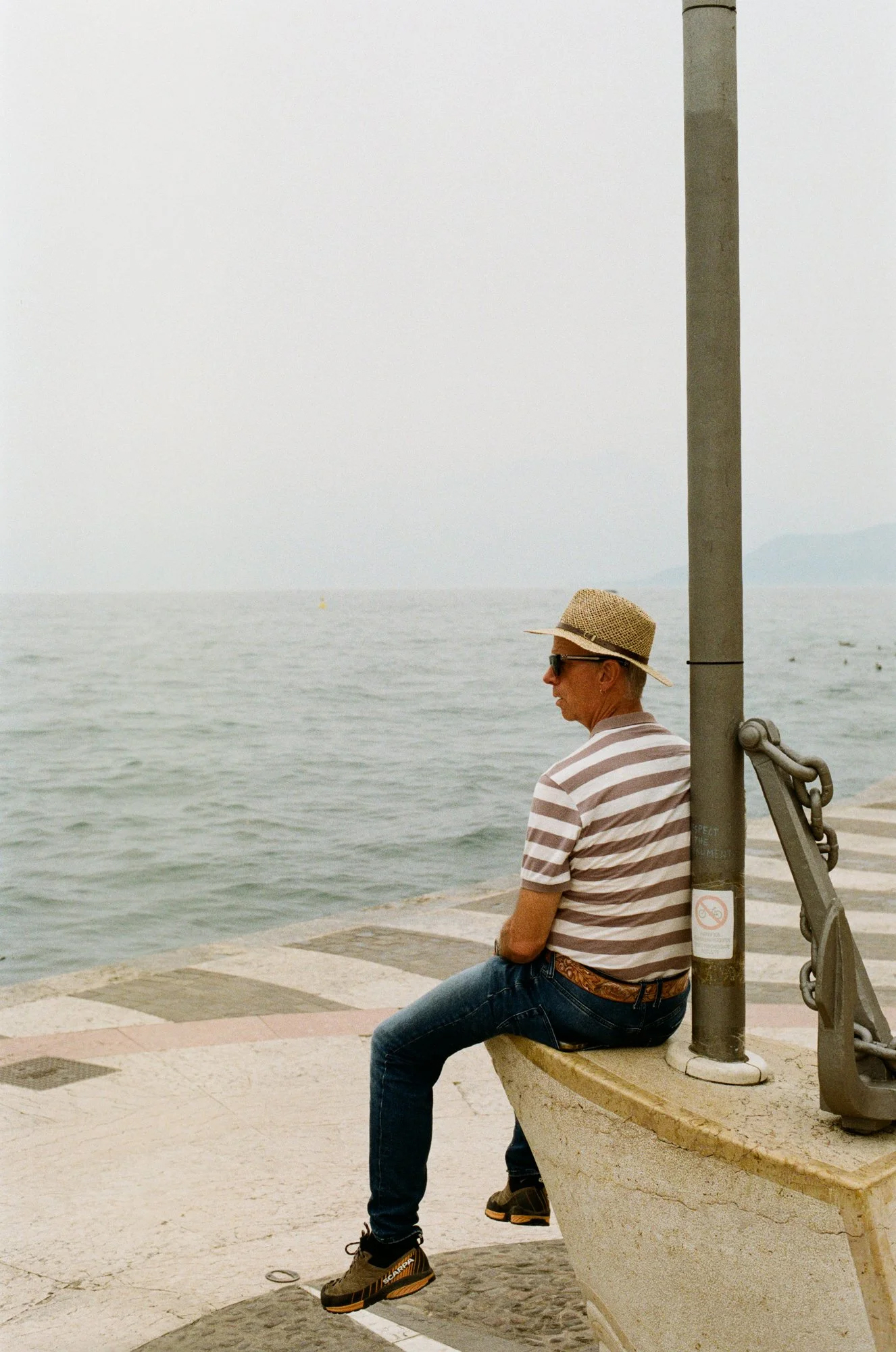 Man in straw hat, striped shirt, and jeans sitting on a pier by the sea.