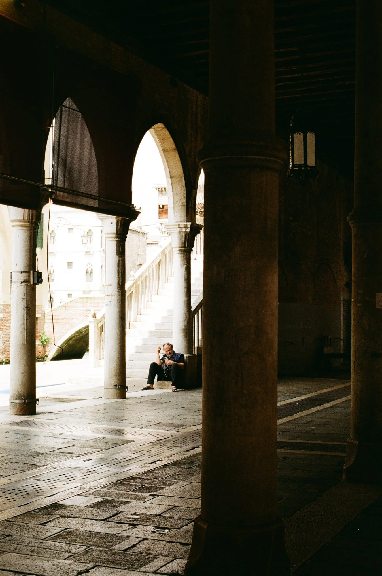 Bearded man sits in shaded stone archway, adjusting headphones, with bright canal background.