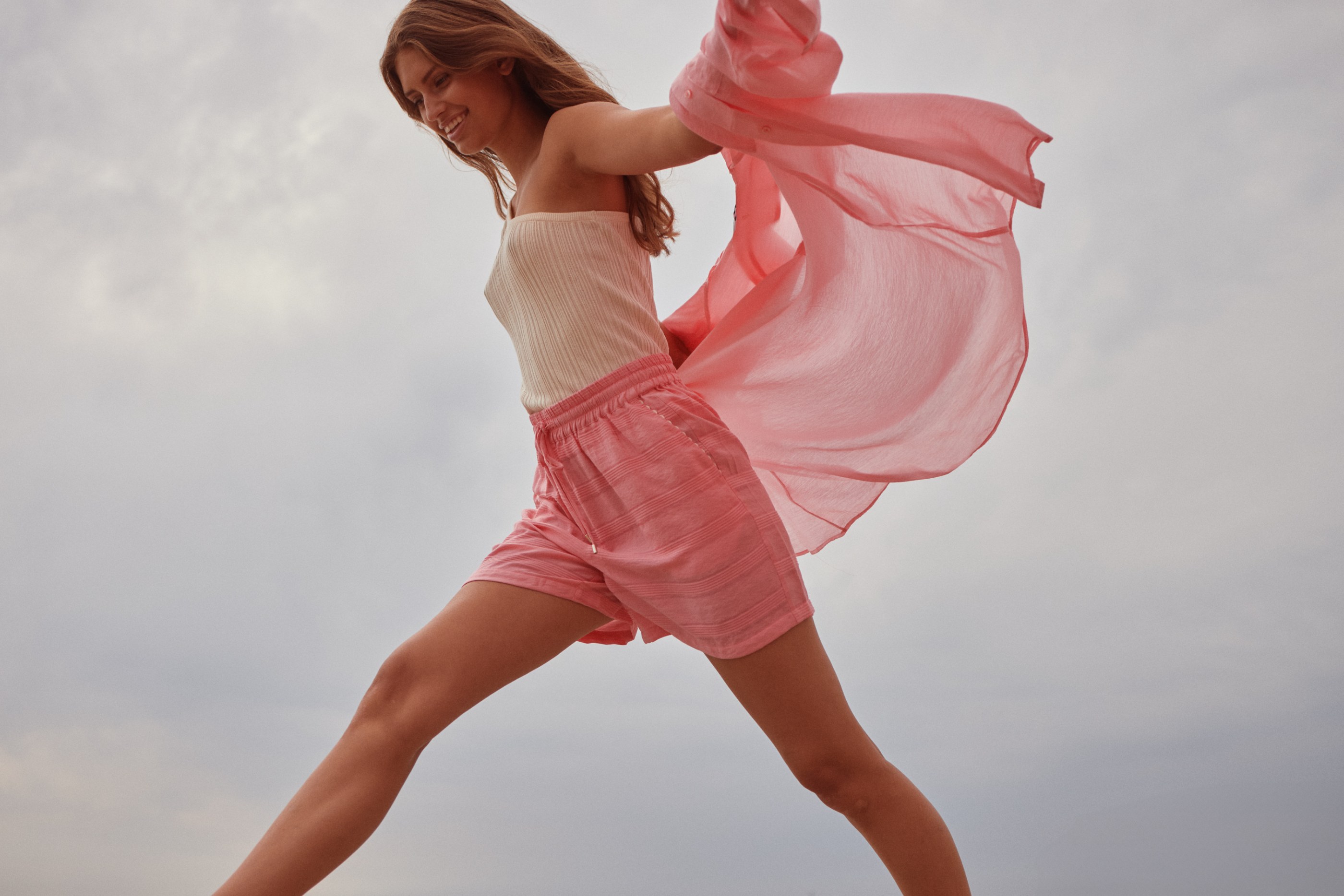 Smiling woman in ribbed top and pink shorts leaps with flowing pink fabric against a cloudy sky.