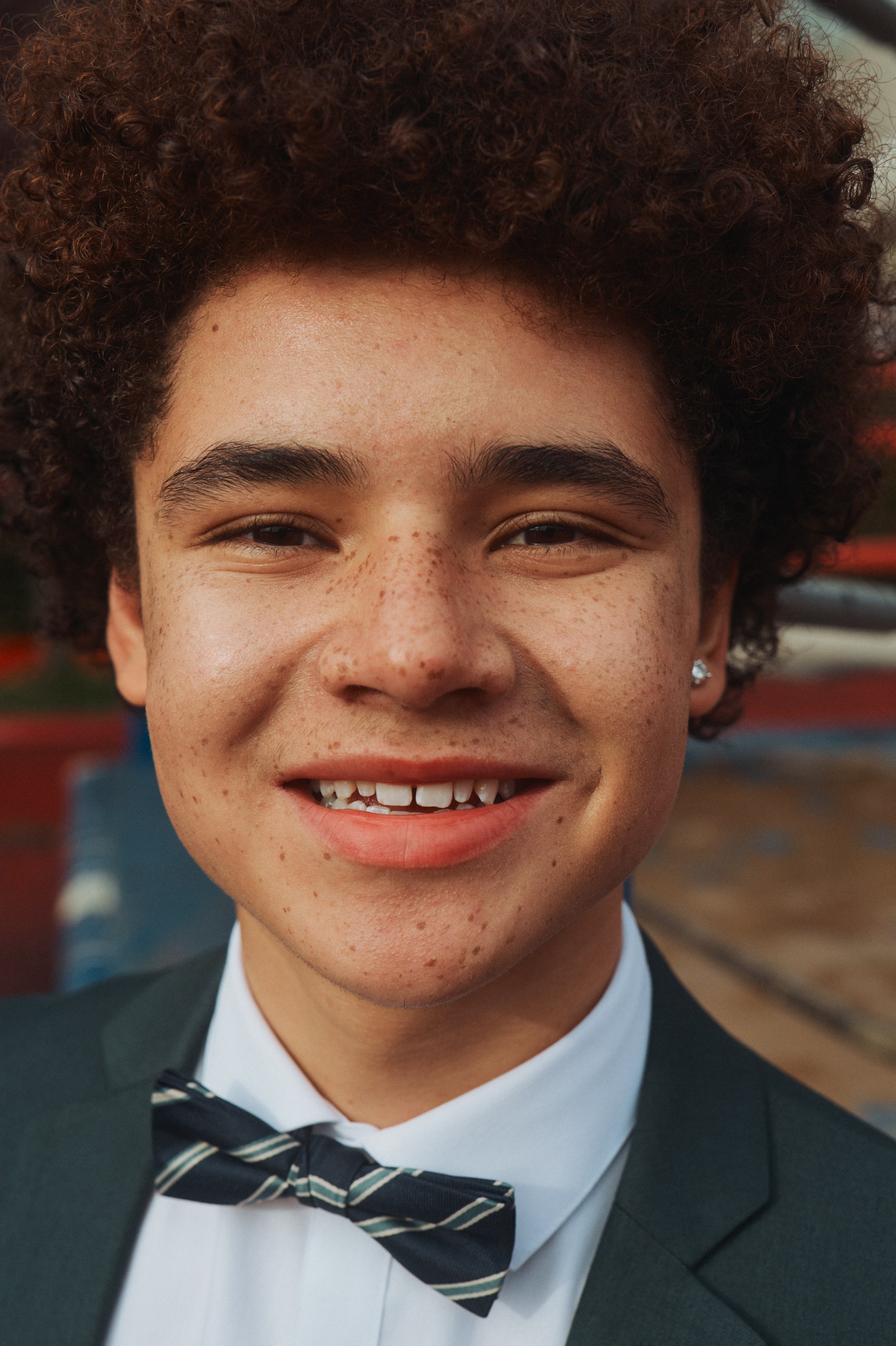 Close-up of a smiling young person with curly hair, freckles, wearing a suit and bow tie.
