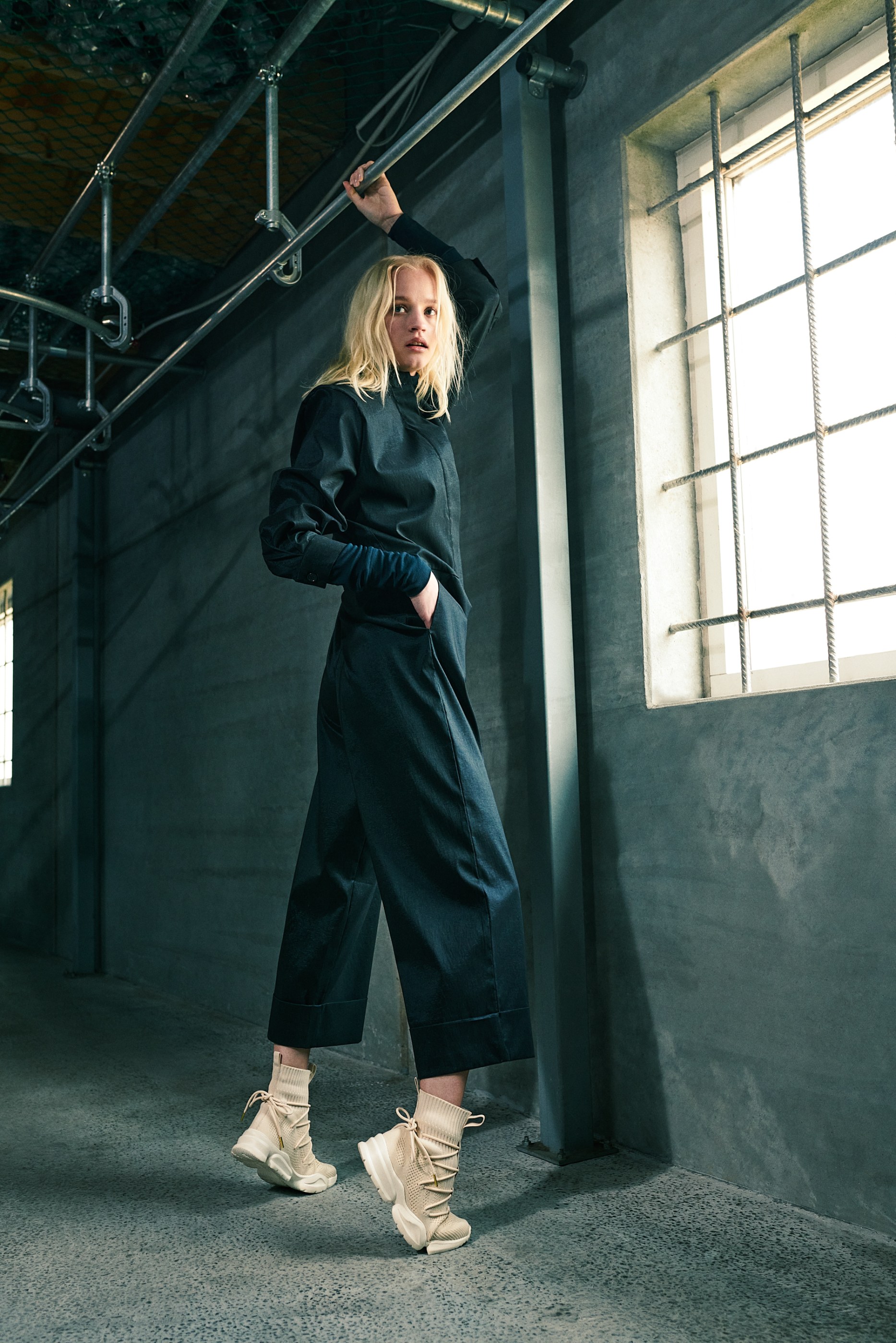 Blonde woman in dark blue outfit and cream sneakers holds overhead pipe in industrial setting.