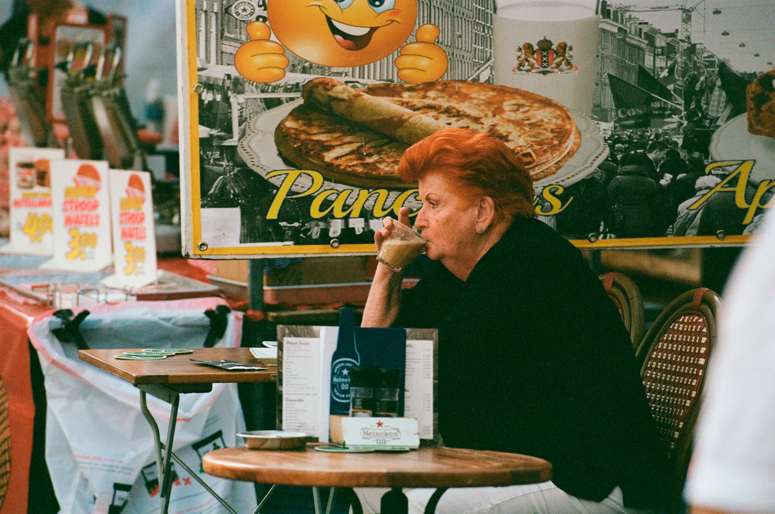 Red-haired woman sips drink at an outdoor cafe table with a large pancake advertisement.