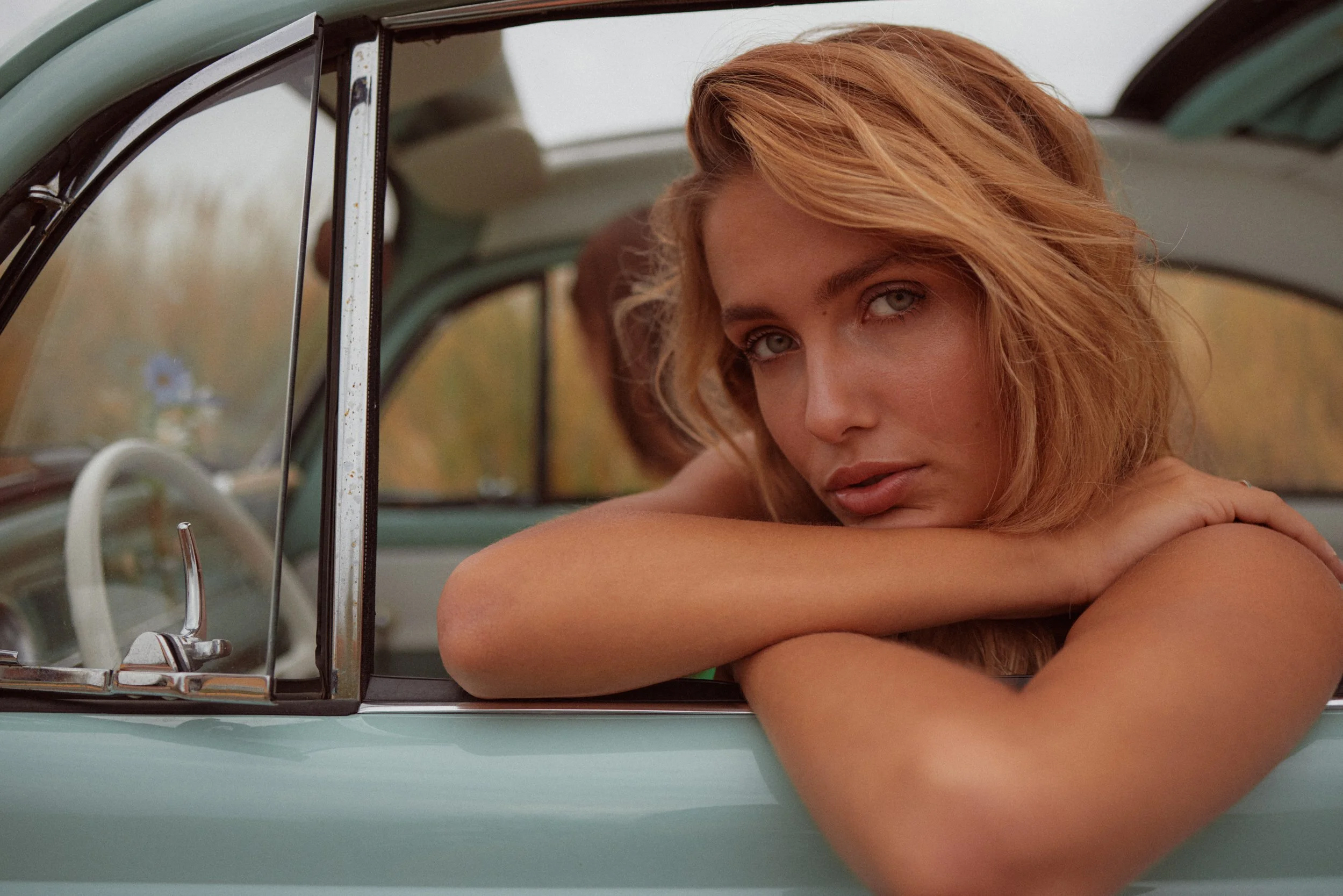 Blonde woman with green eyes leaning on crossed arms out of a vintage teal car.