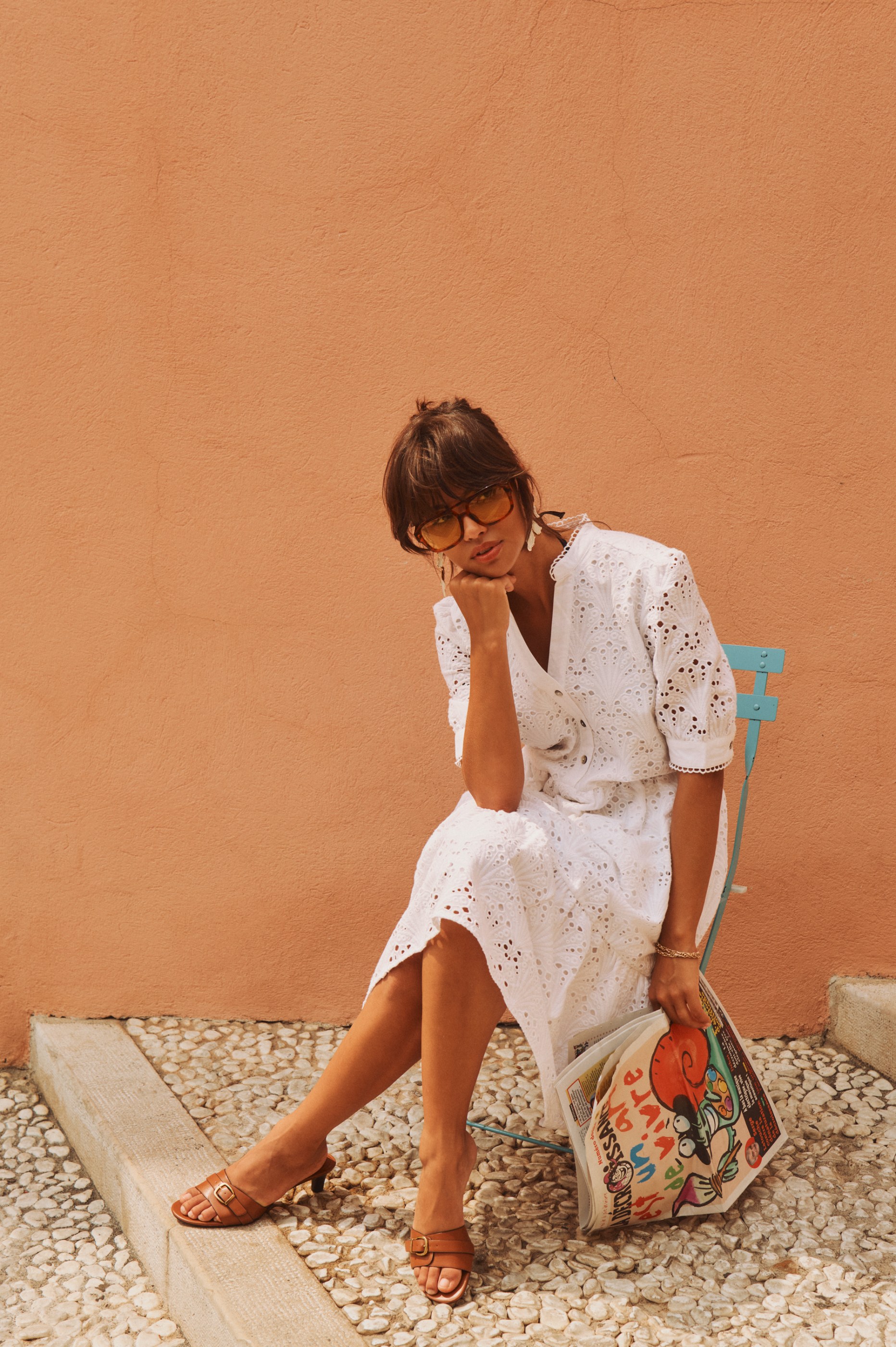Woman in white eyelet dress and sunglasses sits on a blue chair, holding a newspaper.