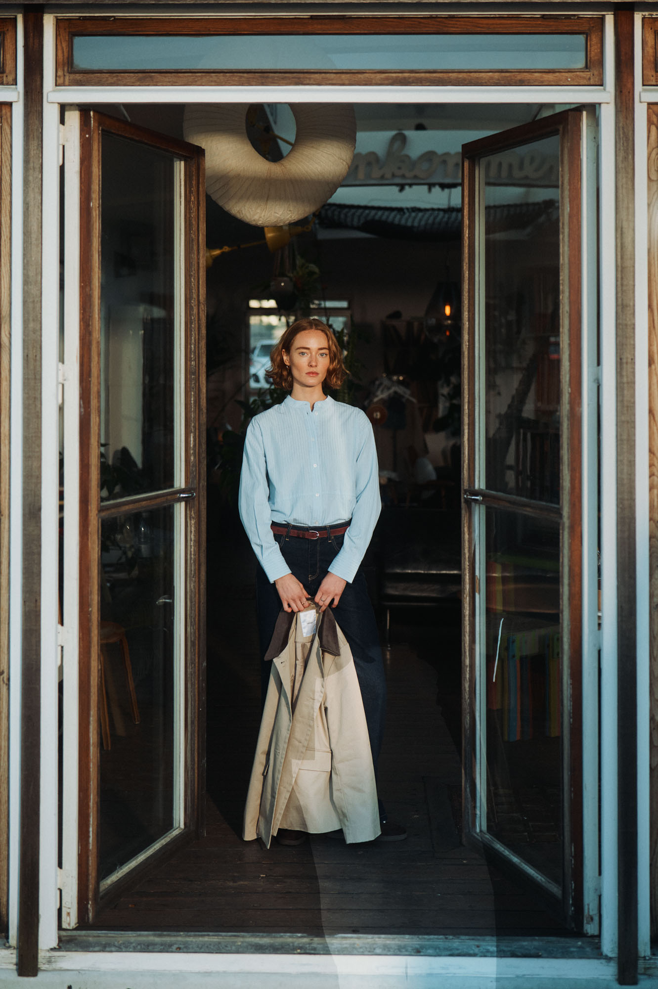 Young woman in a blue shirt and jeans holds a beige coat in a sunlit doorway.