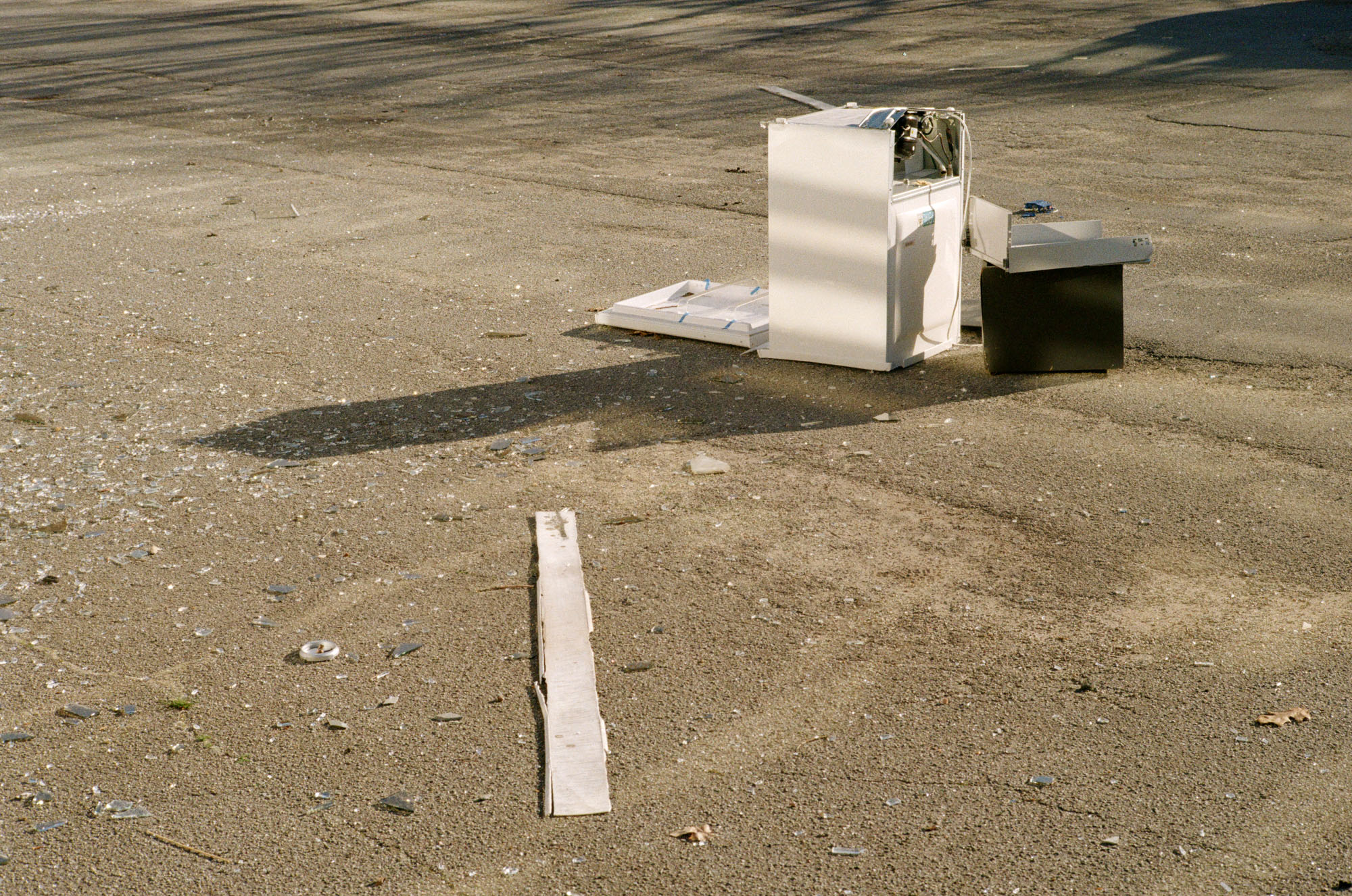 Discarded appliances and broken glass on a sunlit, sandy ground with long shadows.