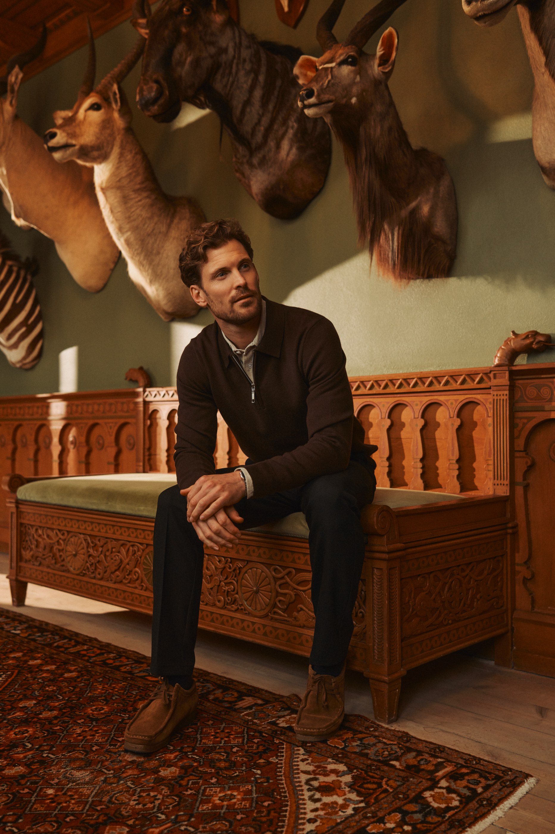 Man in a brown polo shirt sitting on a carved wooden bench surrounded by taxidermy.