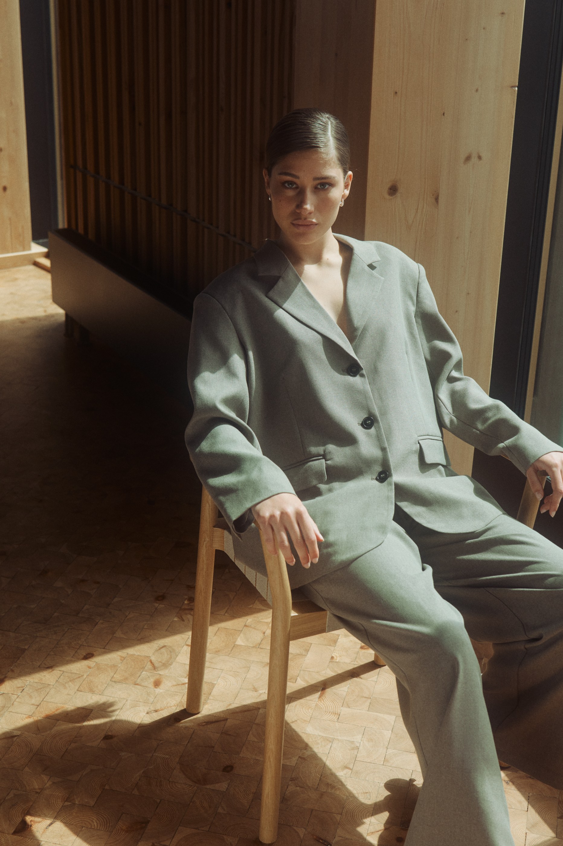 Woman in an oversized grey suit sitting in a sunlit wooden room, looking at the camera.