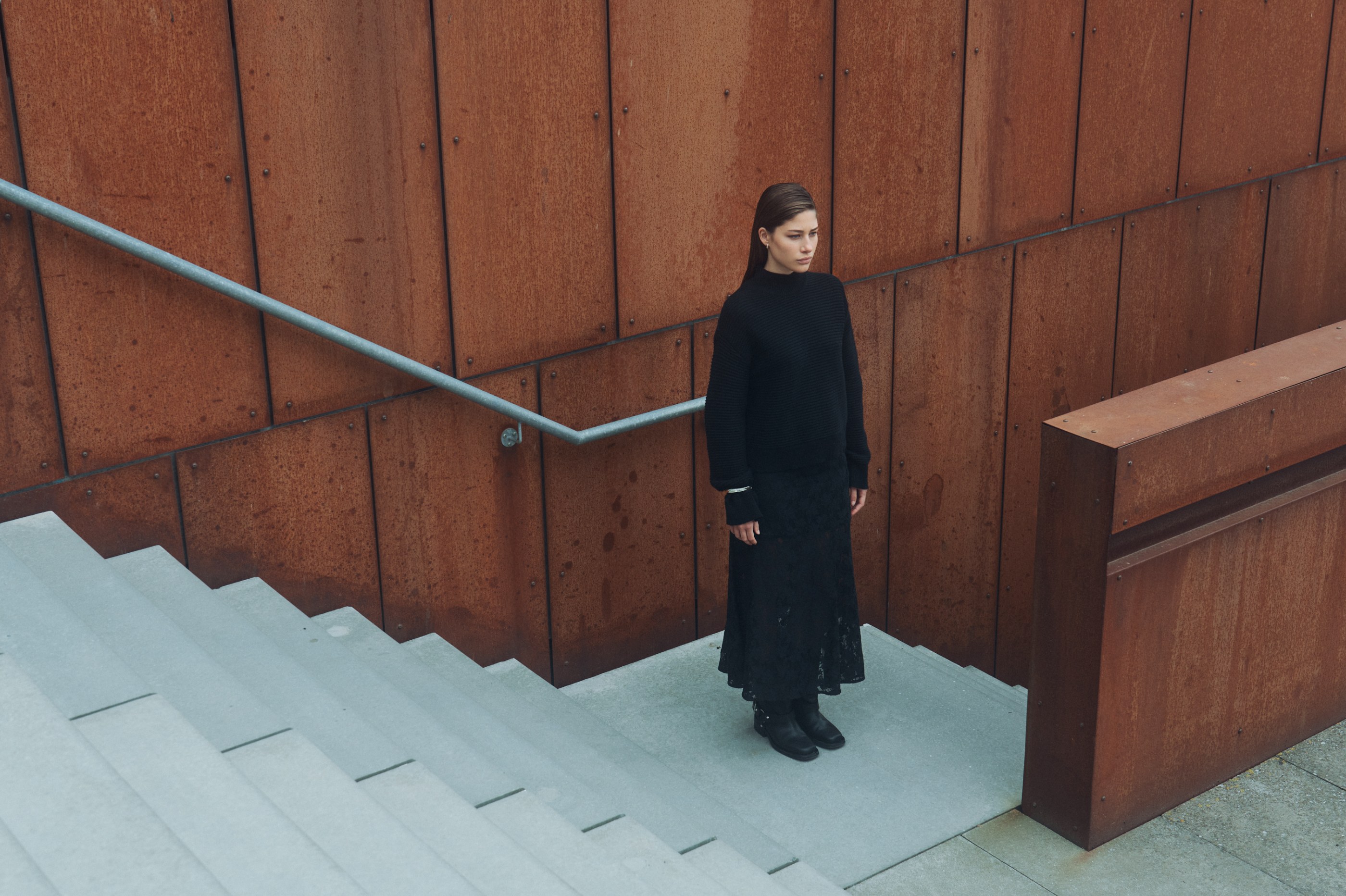 Woman in black knit sweater and lace skirt stands on concrete stairs next to a rusted wall.