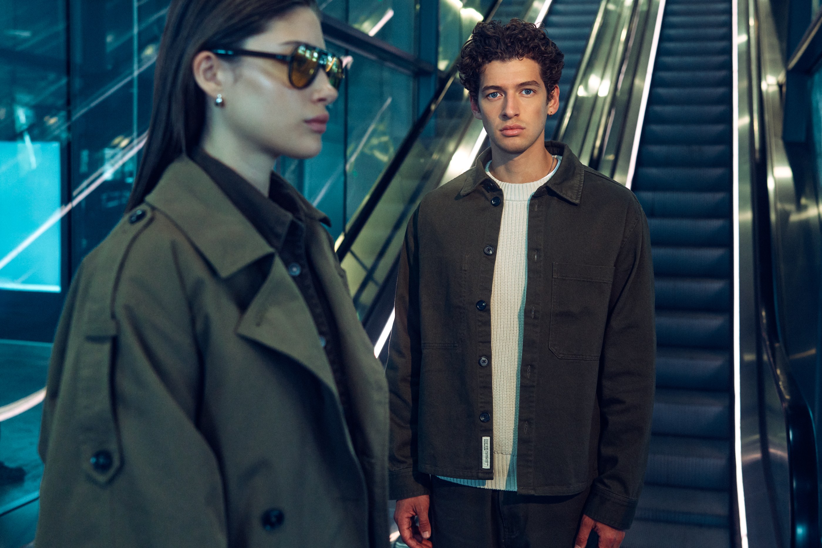 Man in dark jacket and woman in trench coat with sunglasses in escalator setting.