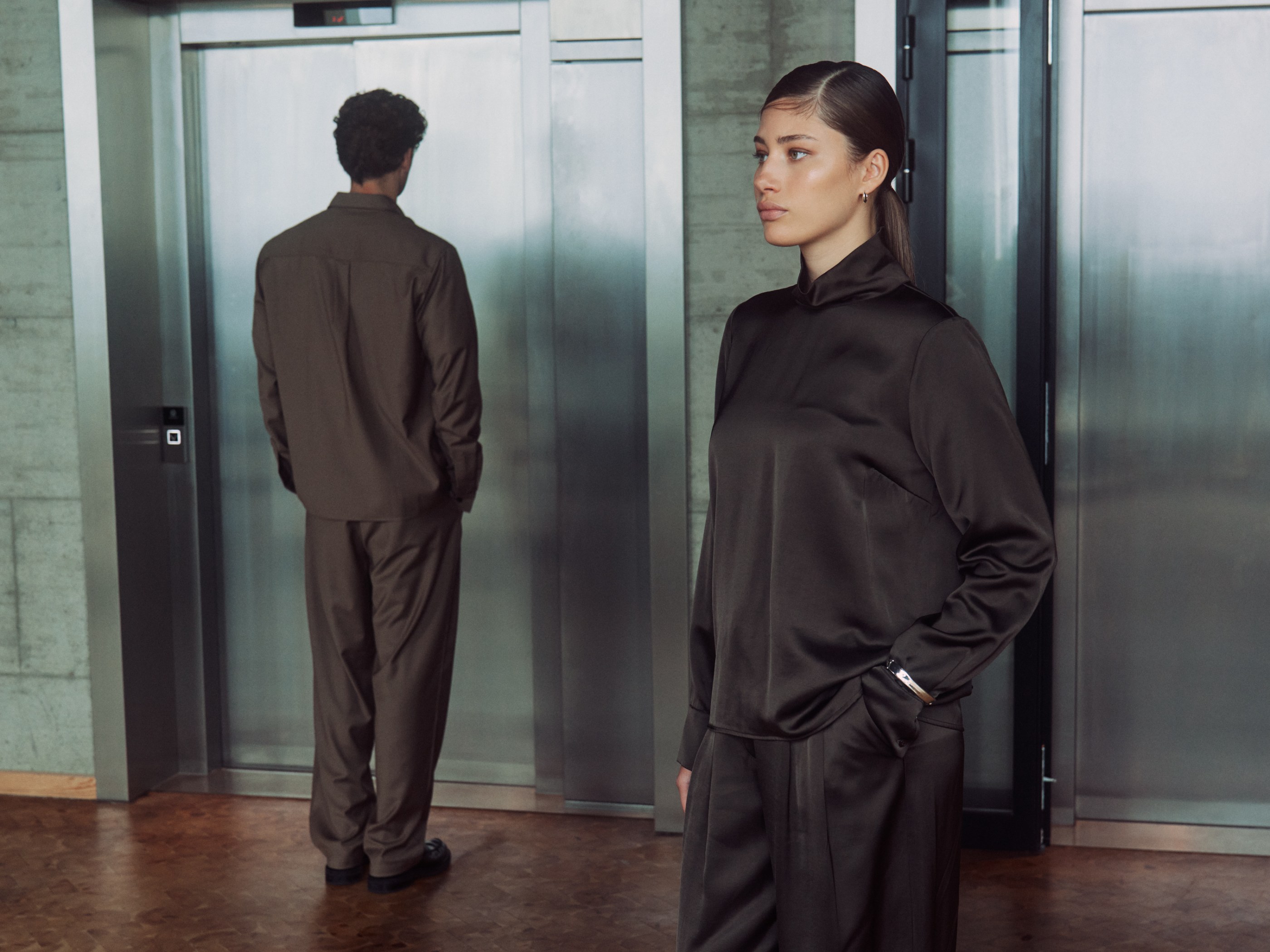 A stylish woman and man in dark brown outfits standing by modern elevators.