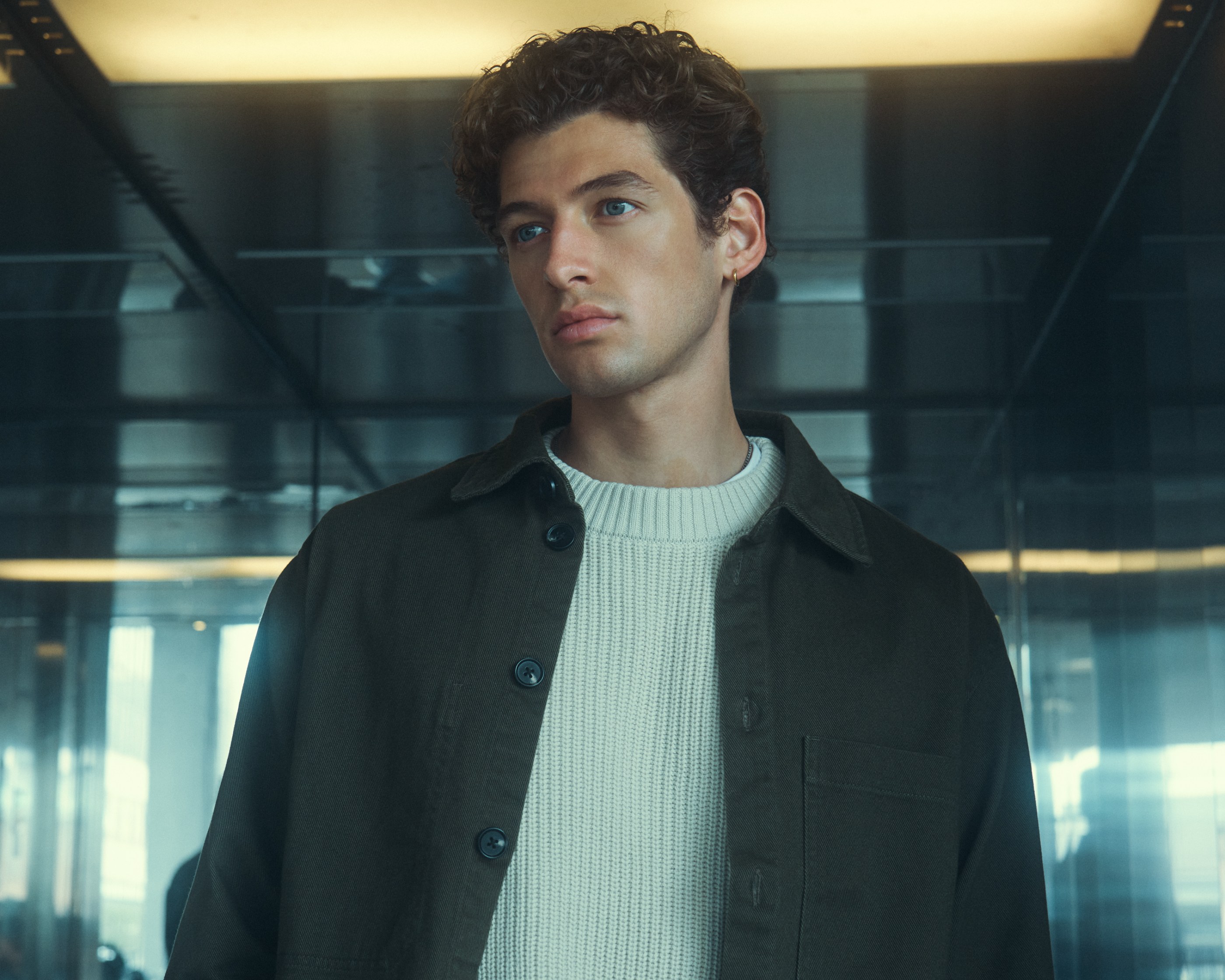 Close-up of a man with curly hair, blue eyes, wearing an olive shacket and cream sweater.