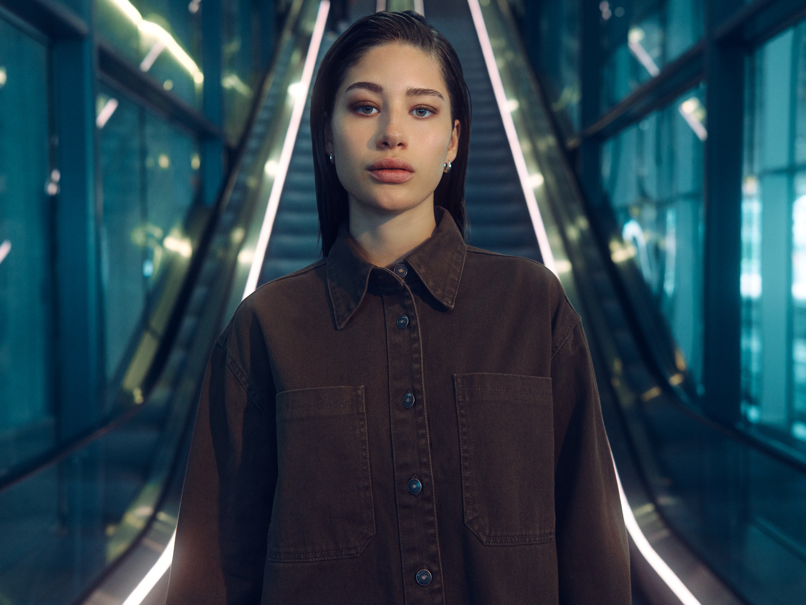 A young woman wearing a brown shirt in front of an illuminated escalator.