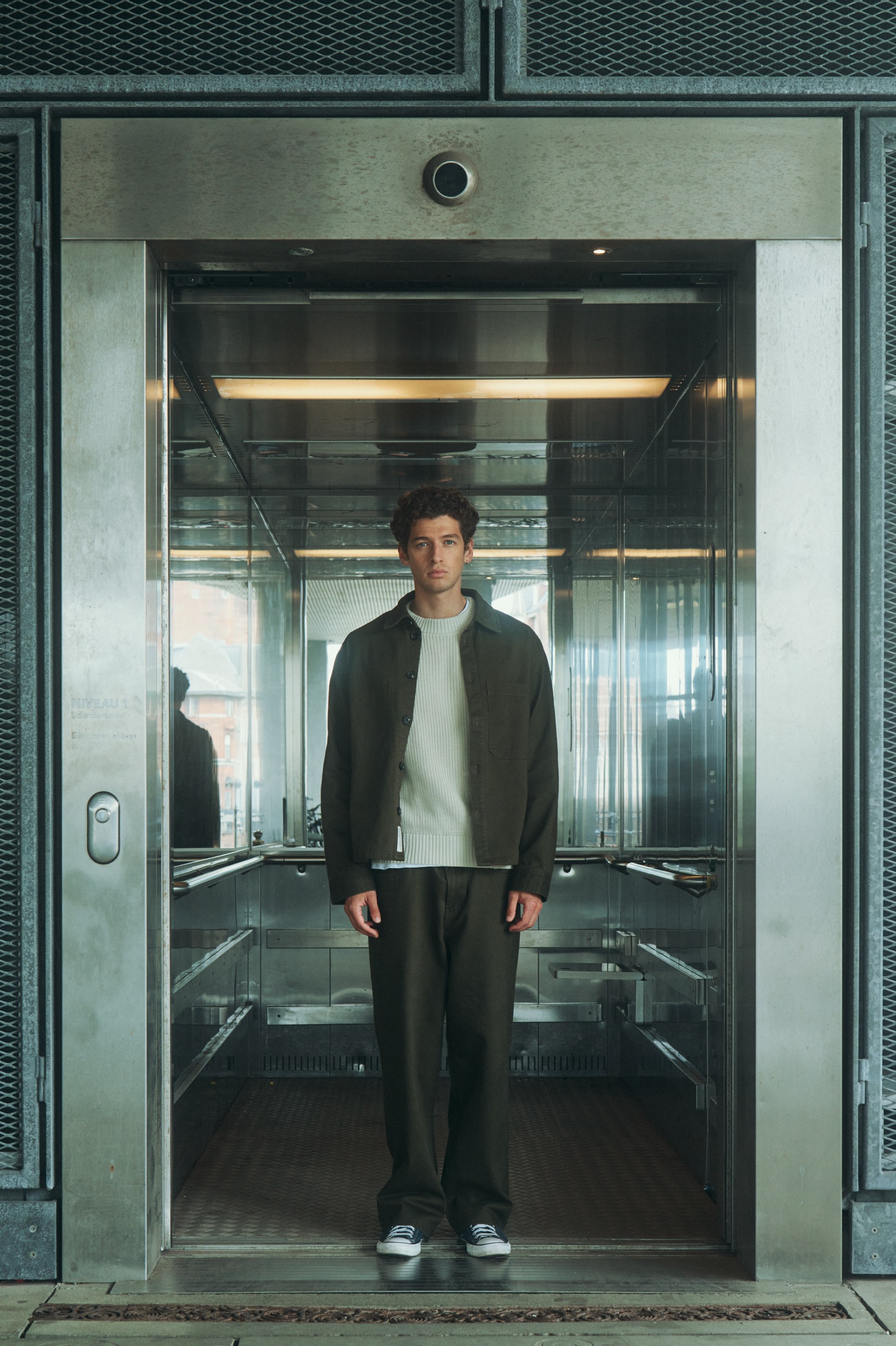 Man in a dark green overshirt and cream sweater stands in a reflective metal elevator.
