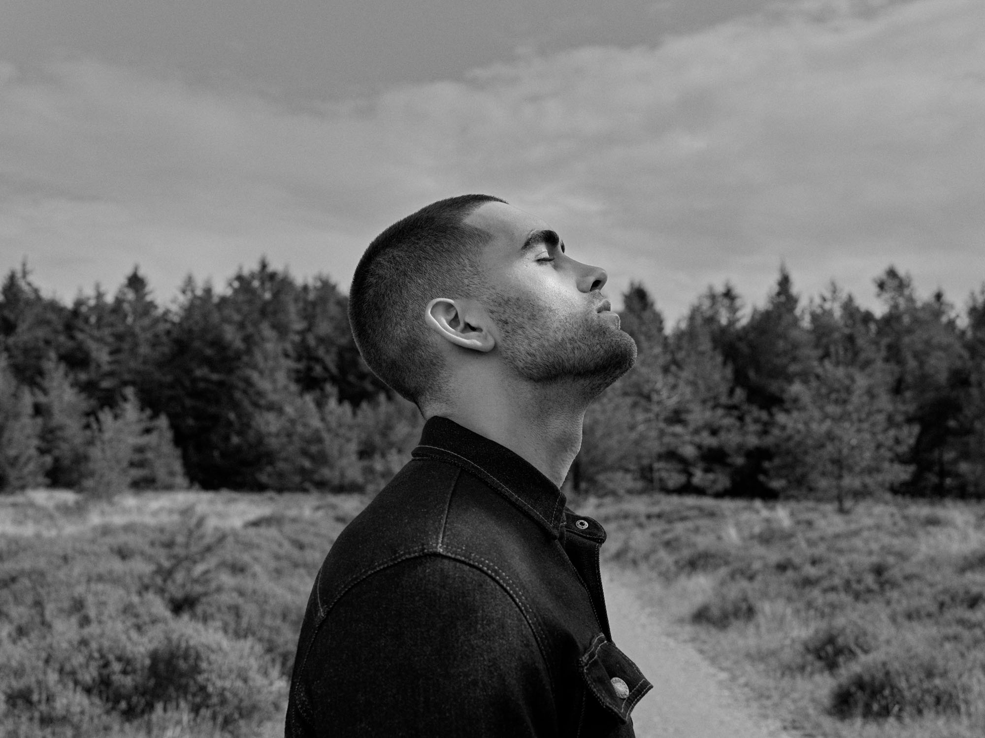 Black and white portrait of a man looking up, eyes closed, wearing a denim jacket outdoors.
