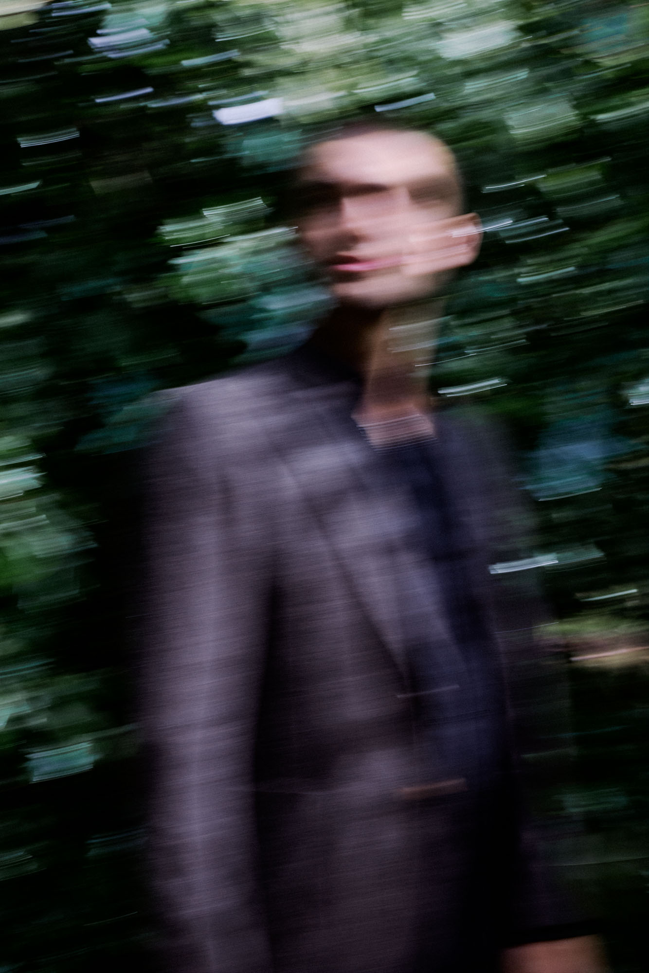 Dynamic portrait of a man in a dark suit with motion blur against green foliage.