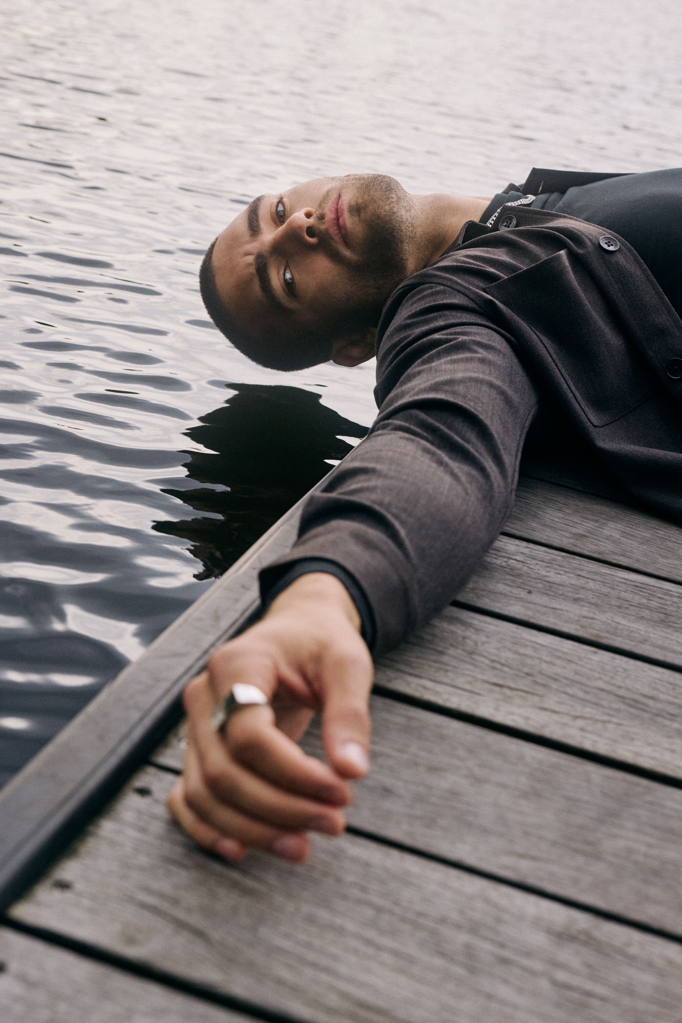 Man in dark jacket lying on wooden dock by water, looking at camera.