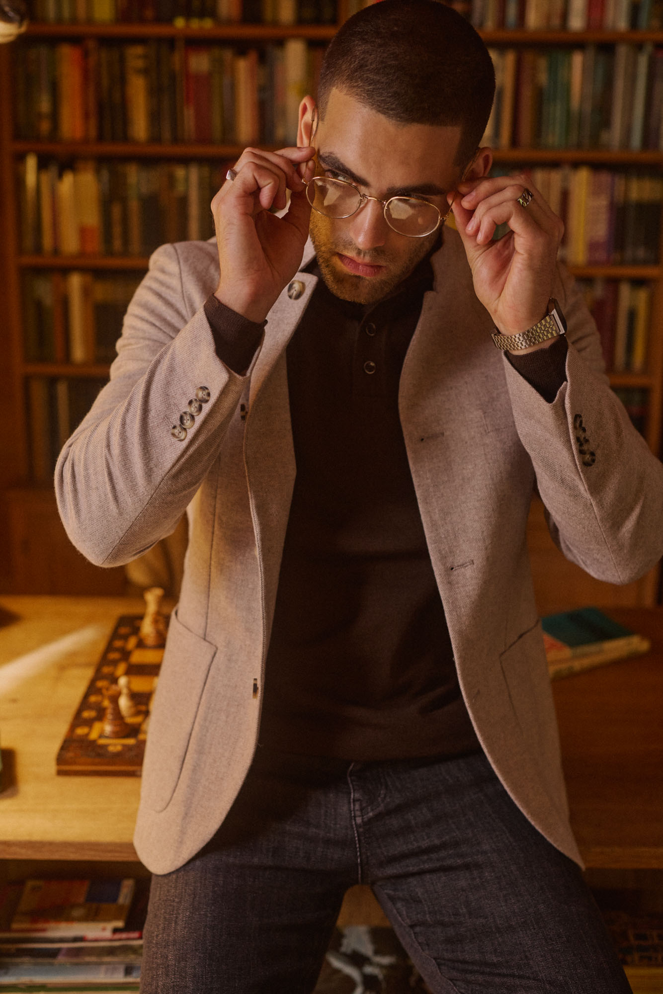 Man adjusting his round glasses in a warm library with bookshelves and a chessboard.