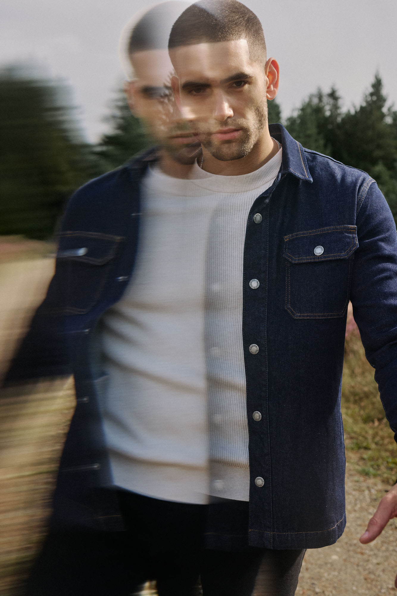 Man with blurred face wearing a dark denim shirt and white long-sleeve tee outdoors with trees.