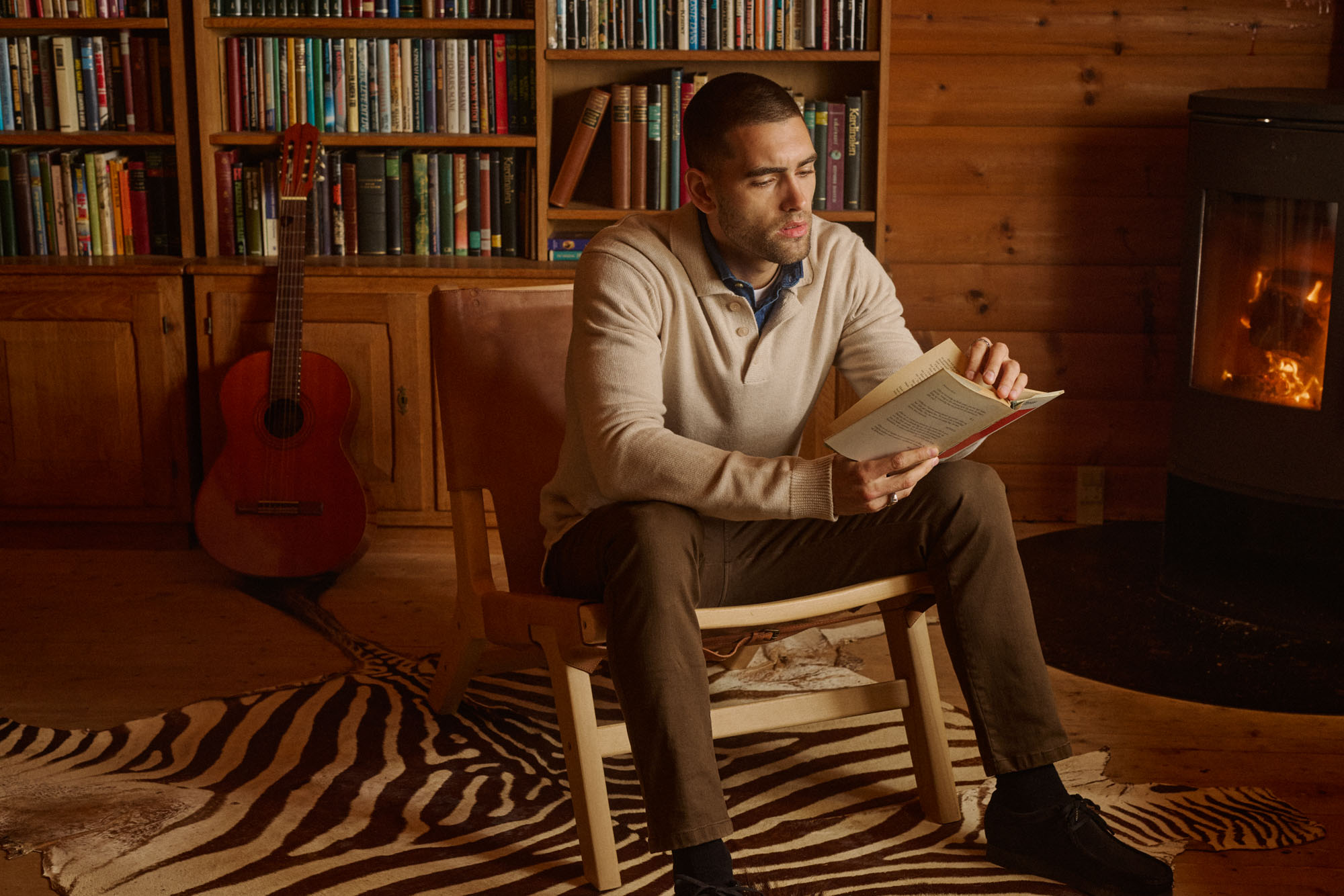 Man reading a book in a cozy rustic cabin with a fireplace, bookshelf, and zebra rug.