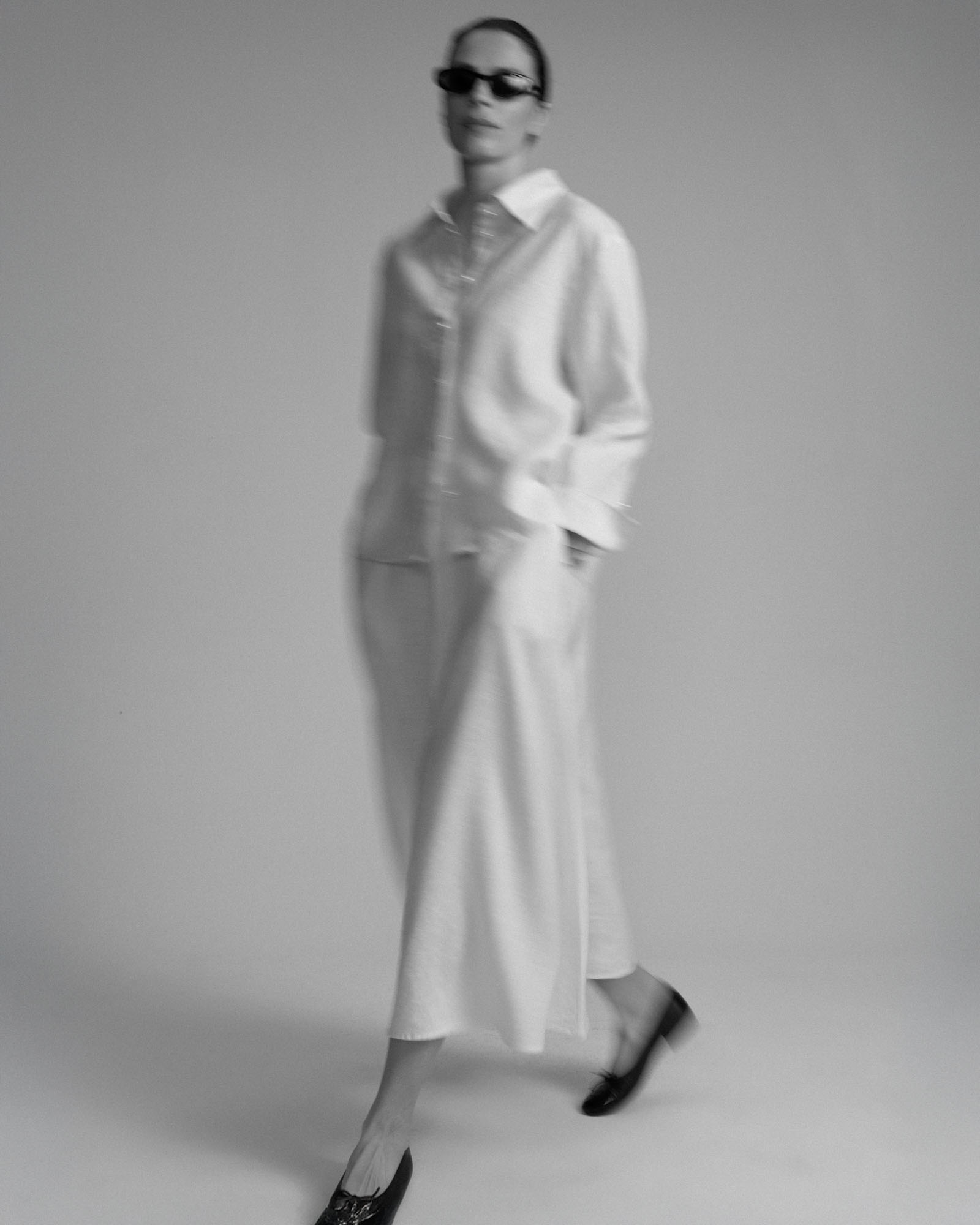 A person in white shirt and trousers, black sunglasses, walks with motion blur in monochrome.