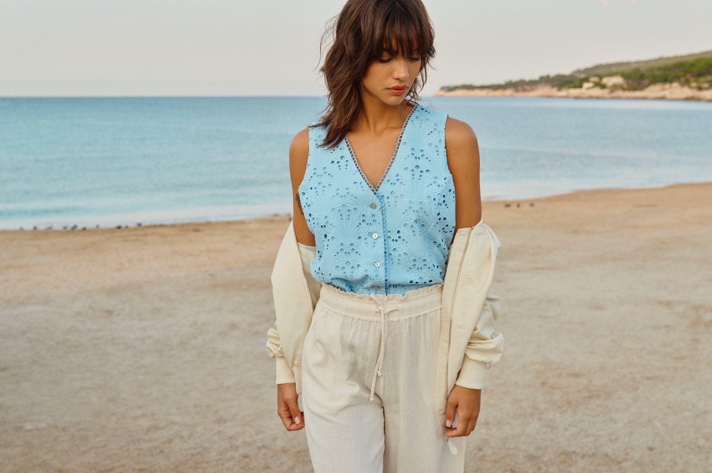 Woman in a light blue eyelet top and cream outfit standing on a sandy beach.