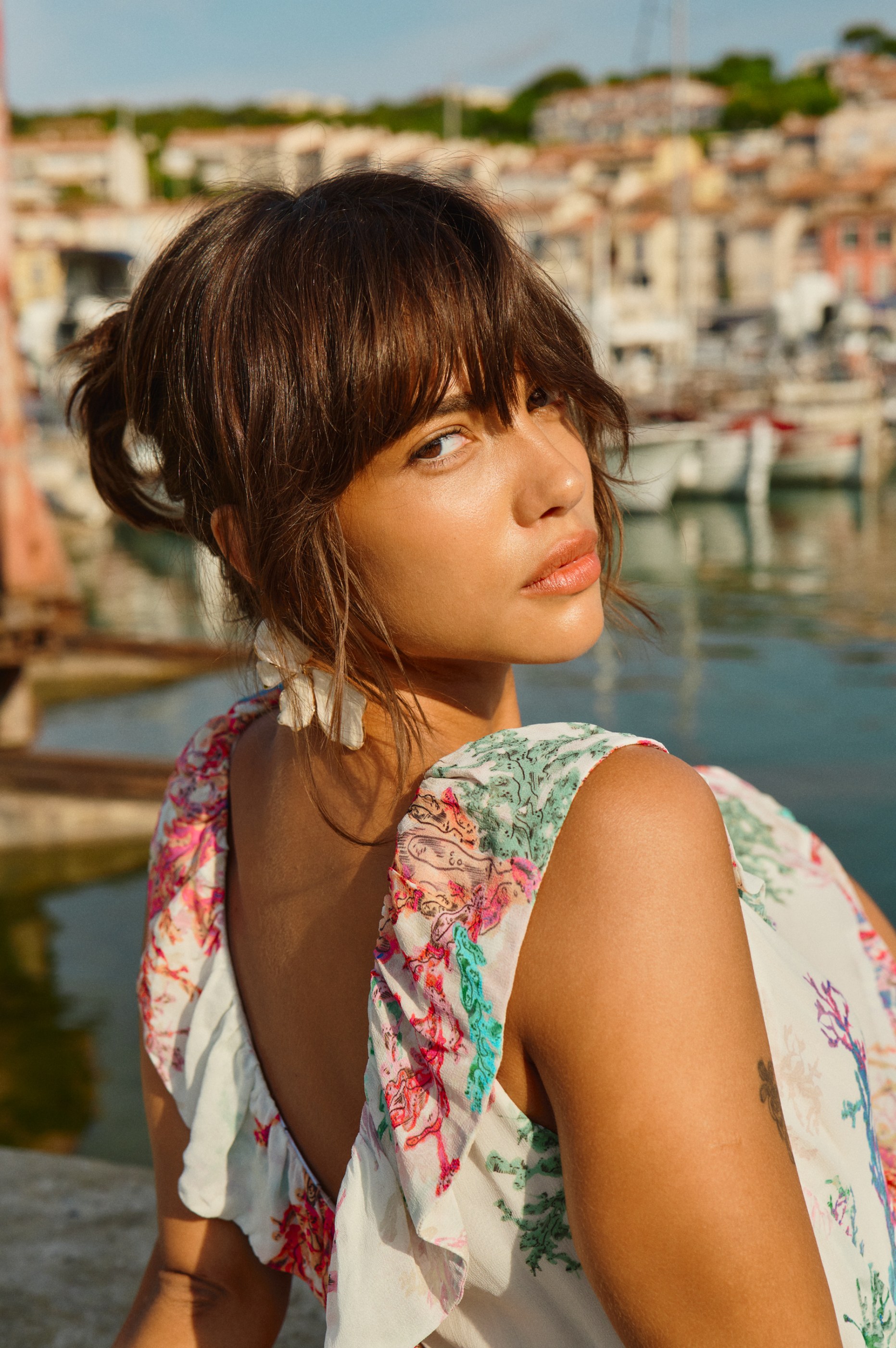 Young woman in patterned dress looking over shoulder in a sunny harbor.