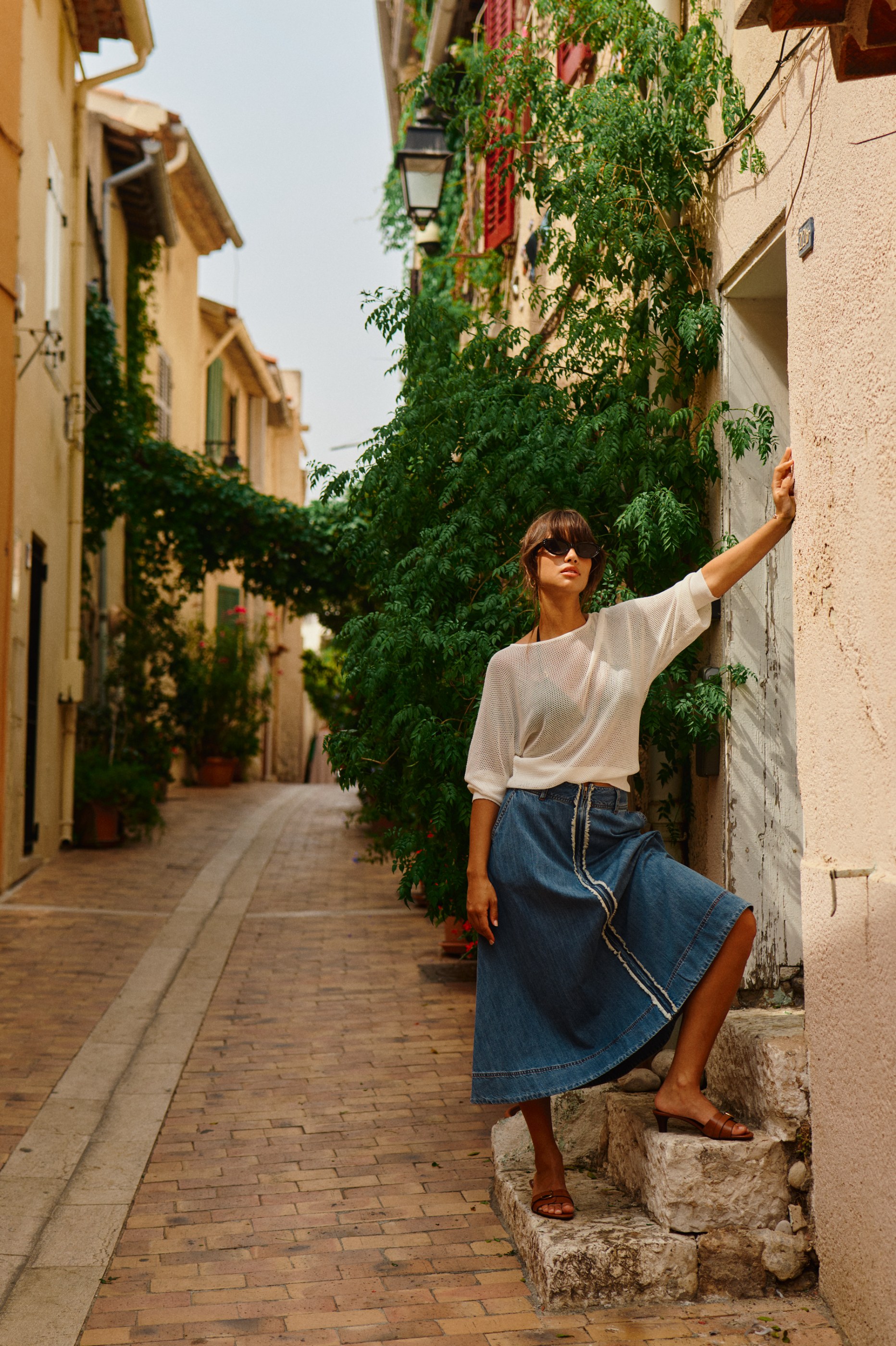 Woman in sunglasses, white top, and denim skirt on stone steps in a sunlit European alley.