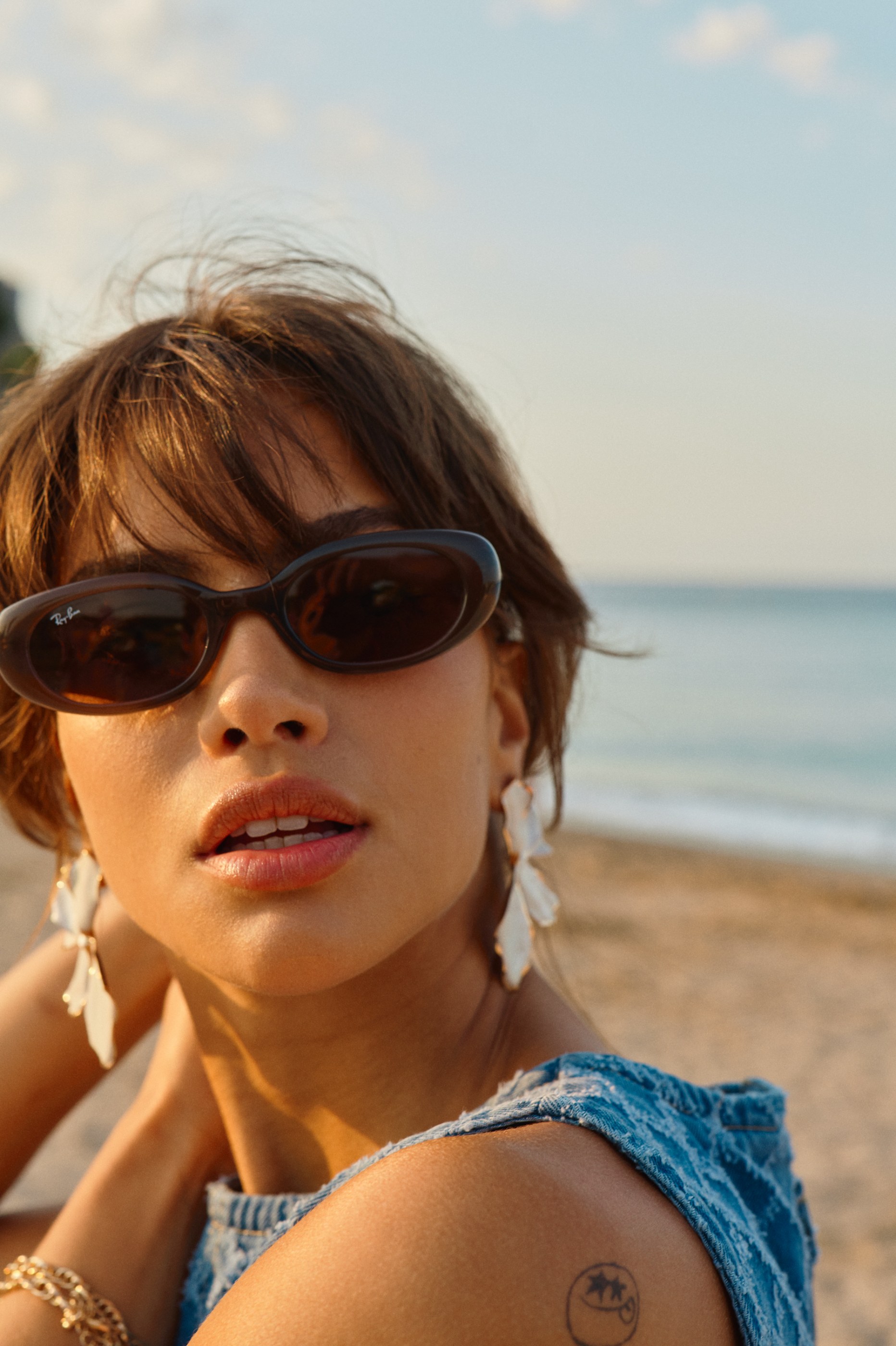 Woman in Ray-Ban sunglasses and shell earrings on a beach at sunset.