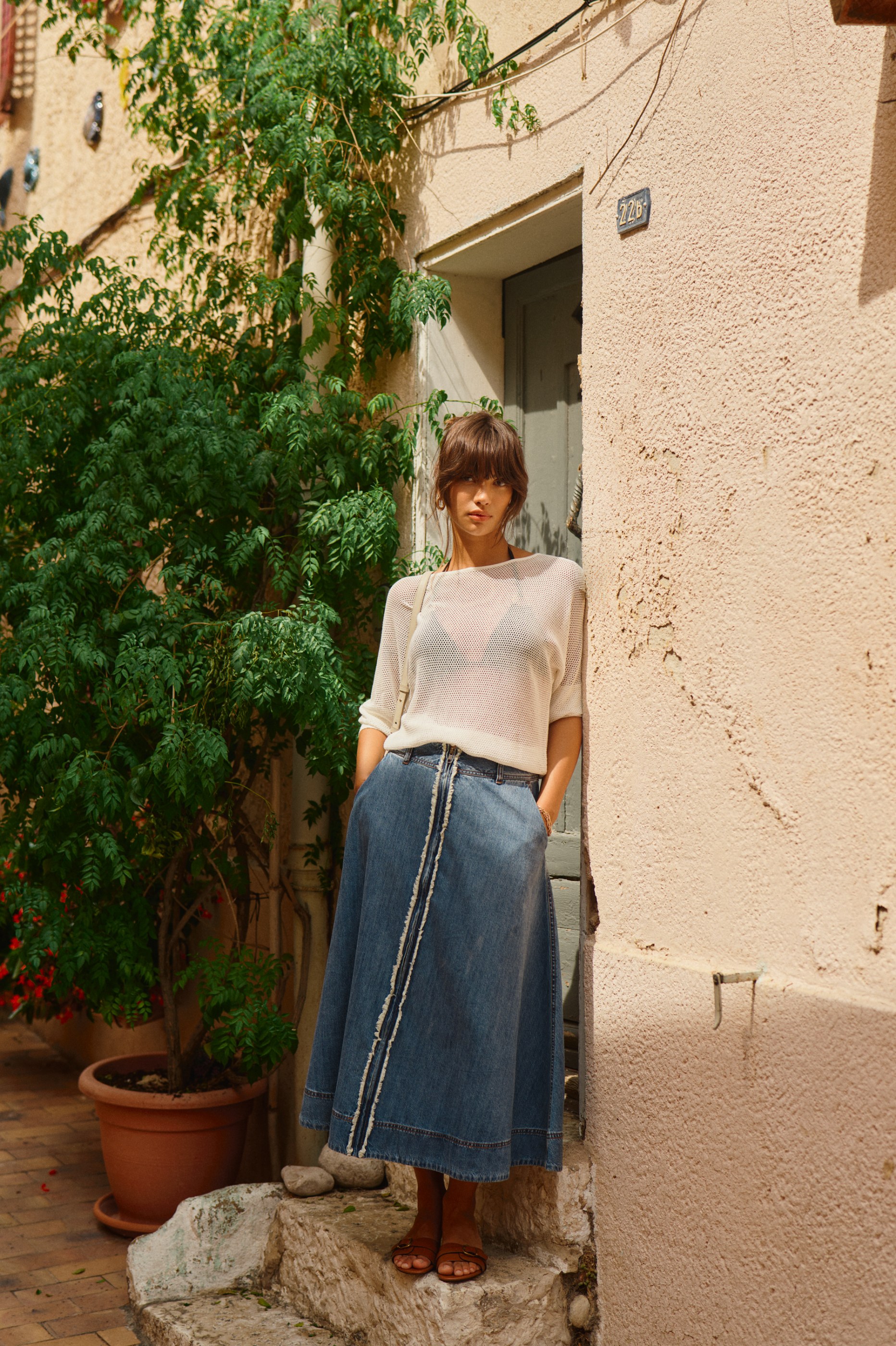 Woman in white knit top and distressed denim maxi skirt standing in a sunlit alley.