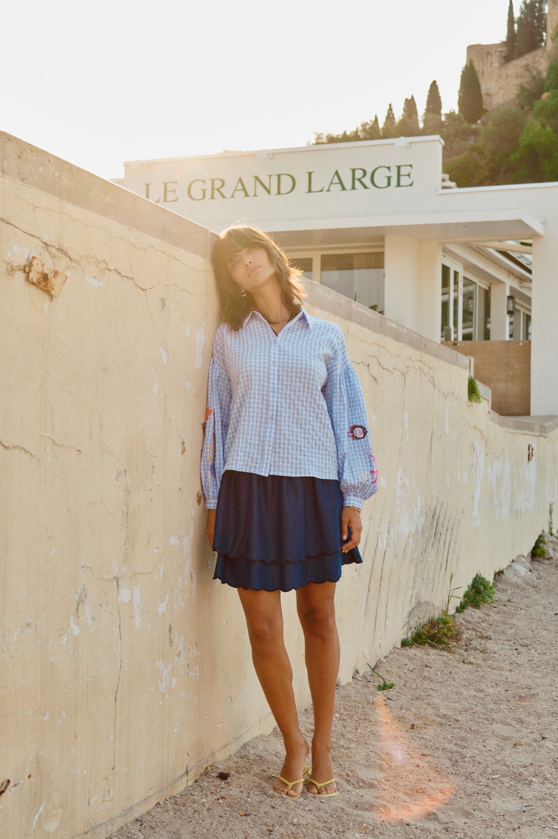 Woman in gingham shirt and navy skirt leans on wall, "LE GRAND LARGE" building, golden hour.