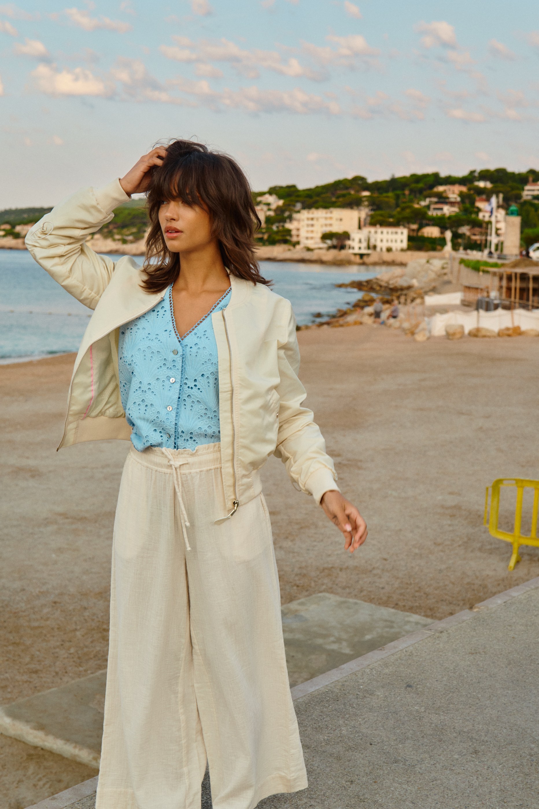 Woman in blue blouse, cream jacket, and beige pants on a sandy beach with a coastal town.