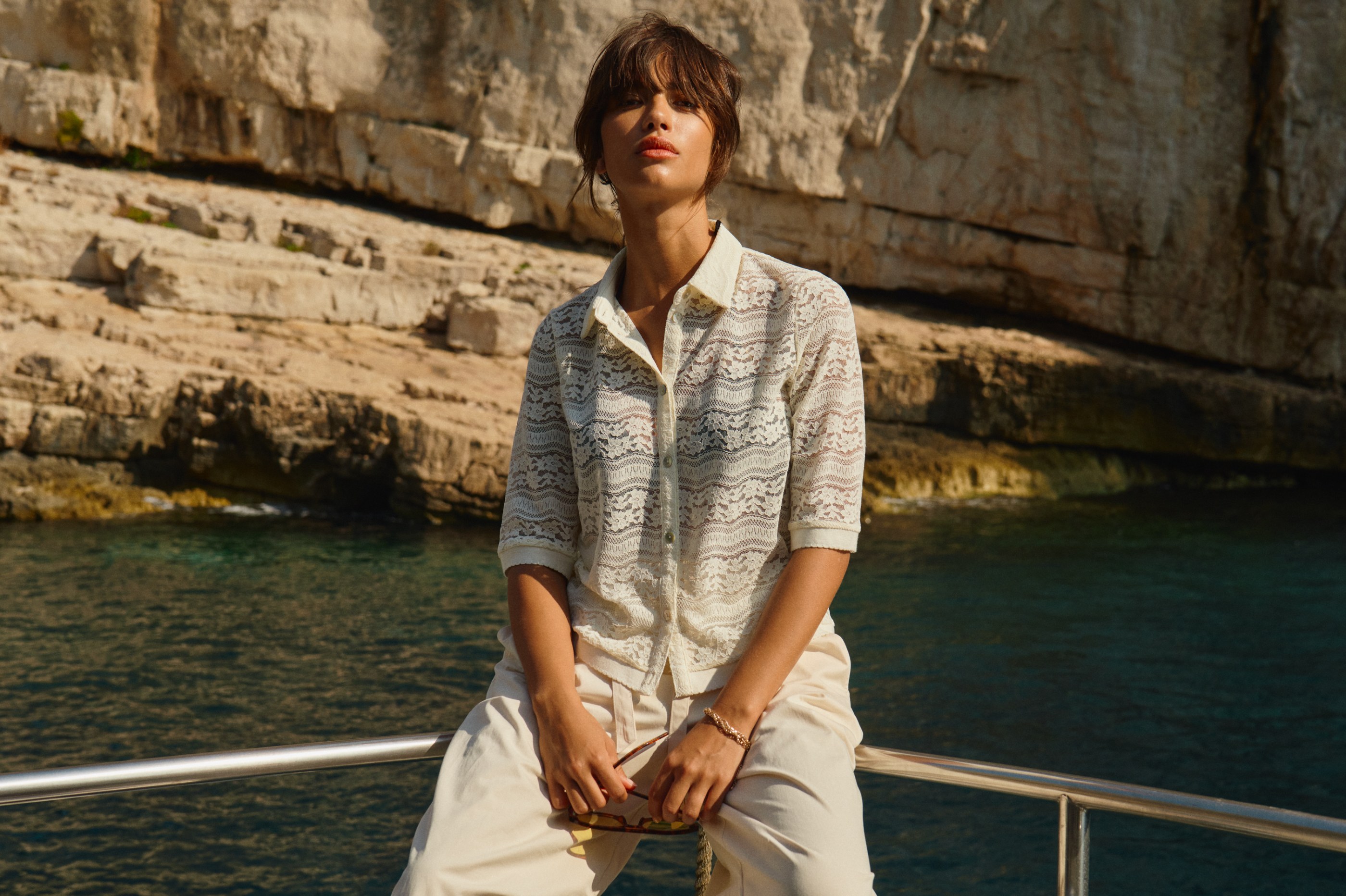 Woman in white lace shirt sitting on boat railing by sunny cliffside and blue water.
