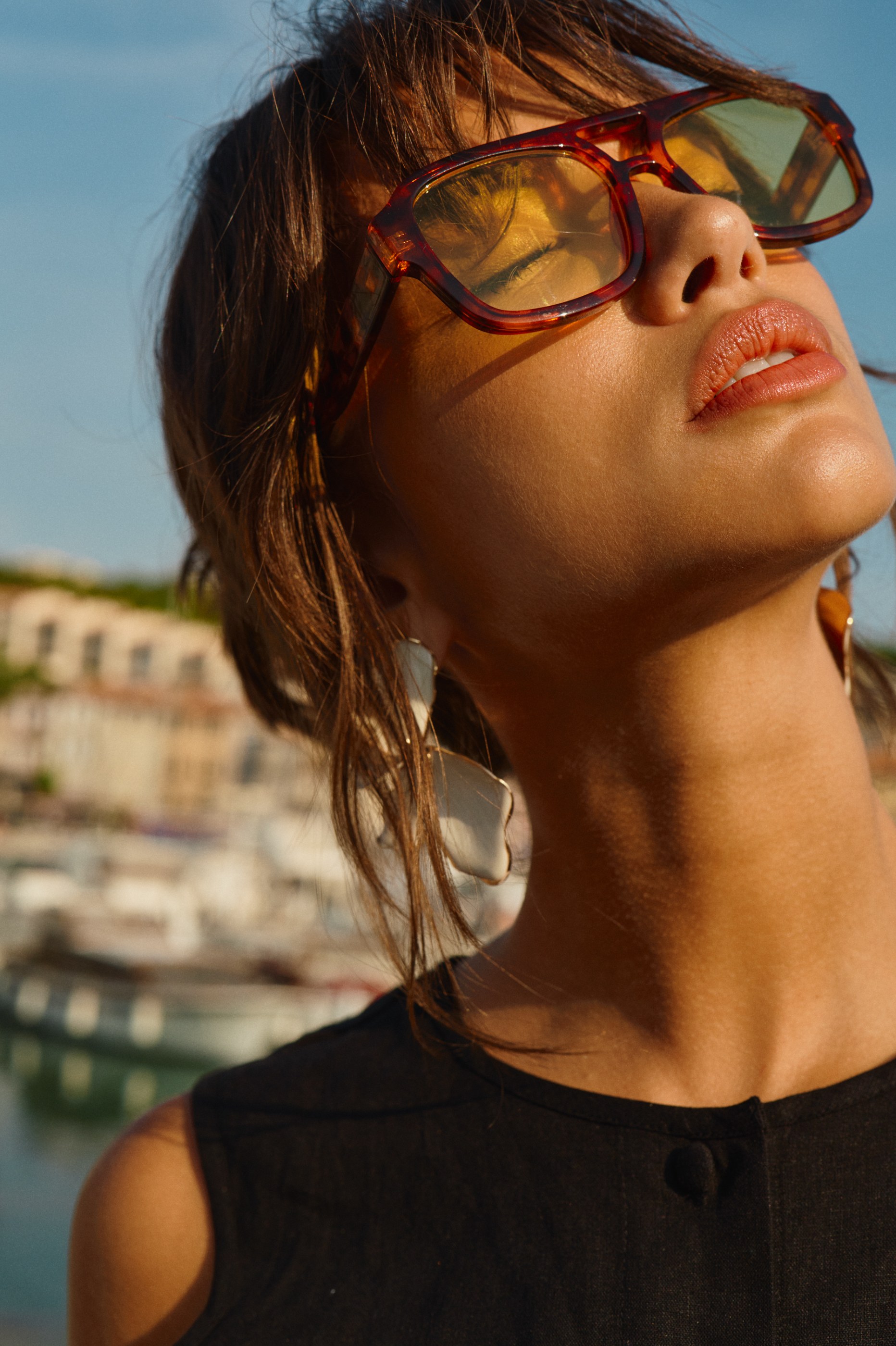 Woman wearing tortoise-shell sunglasses and a black top, looking up in warm sunlight.