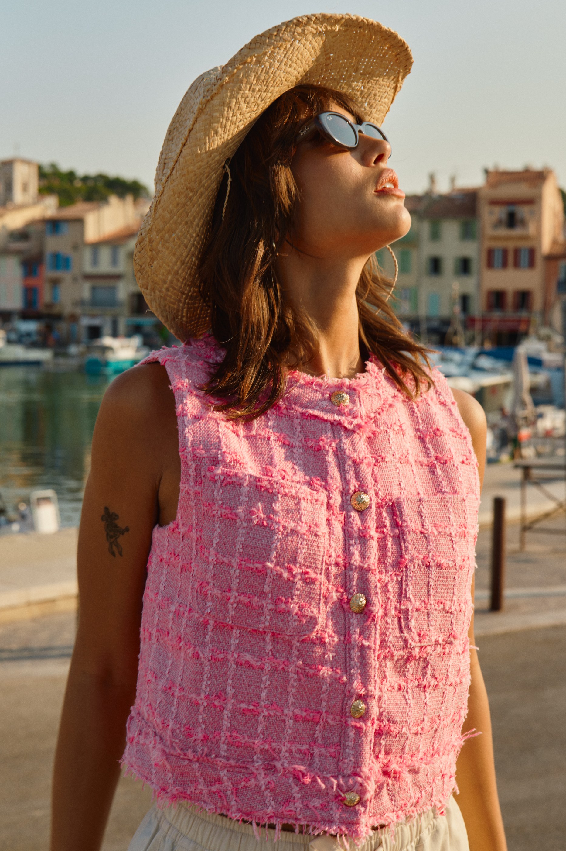Woman in straw hat, Ray-Ban sunglasses, pink top, looking up at sunny harbor.