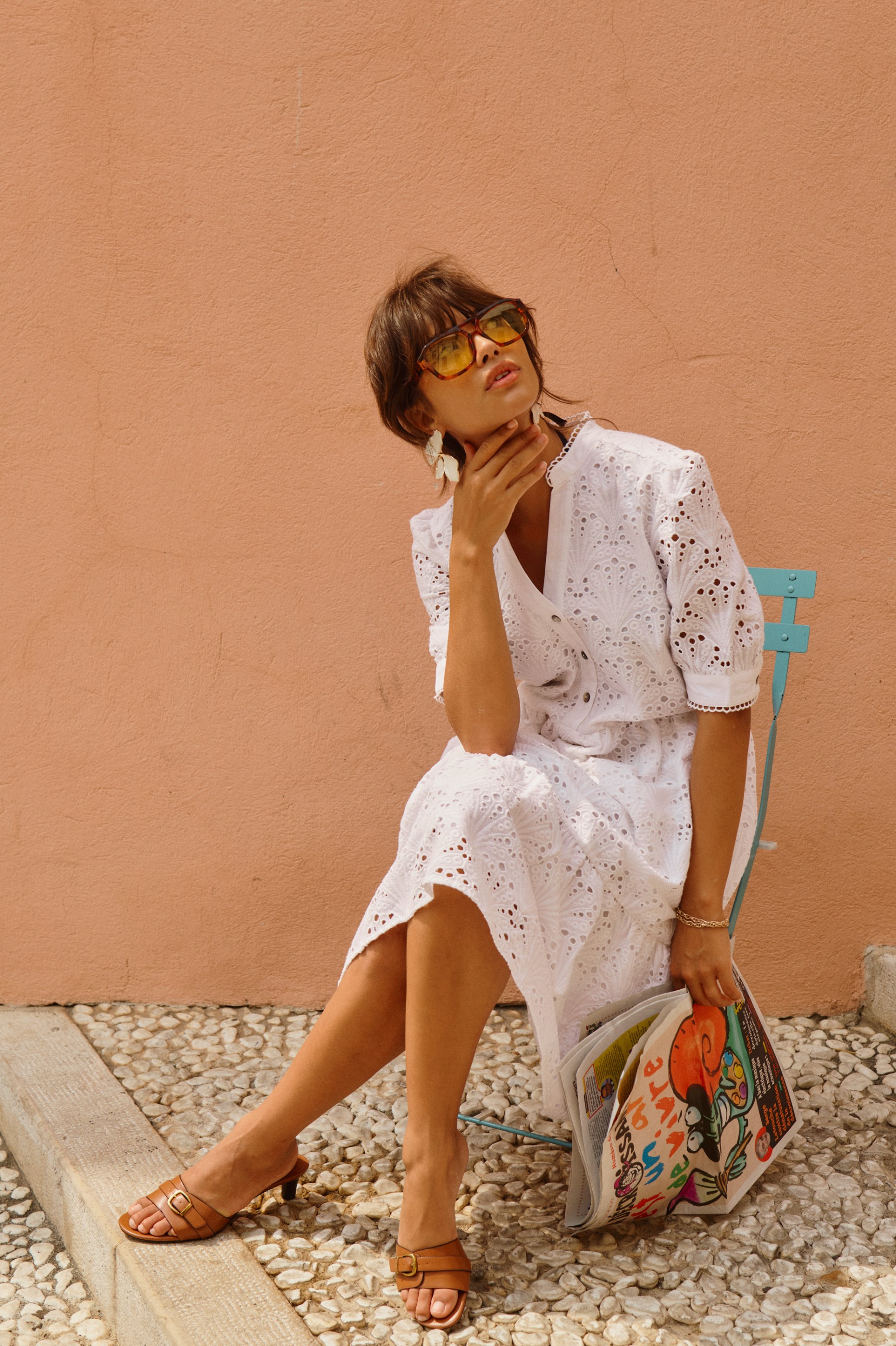Stylish woman in white eyelet dress, yellow sunglasses, holding a newspaper, by a peach wall.