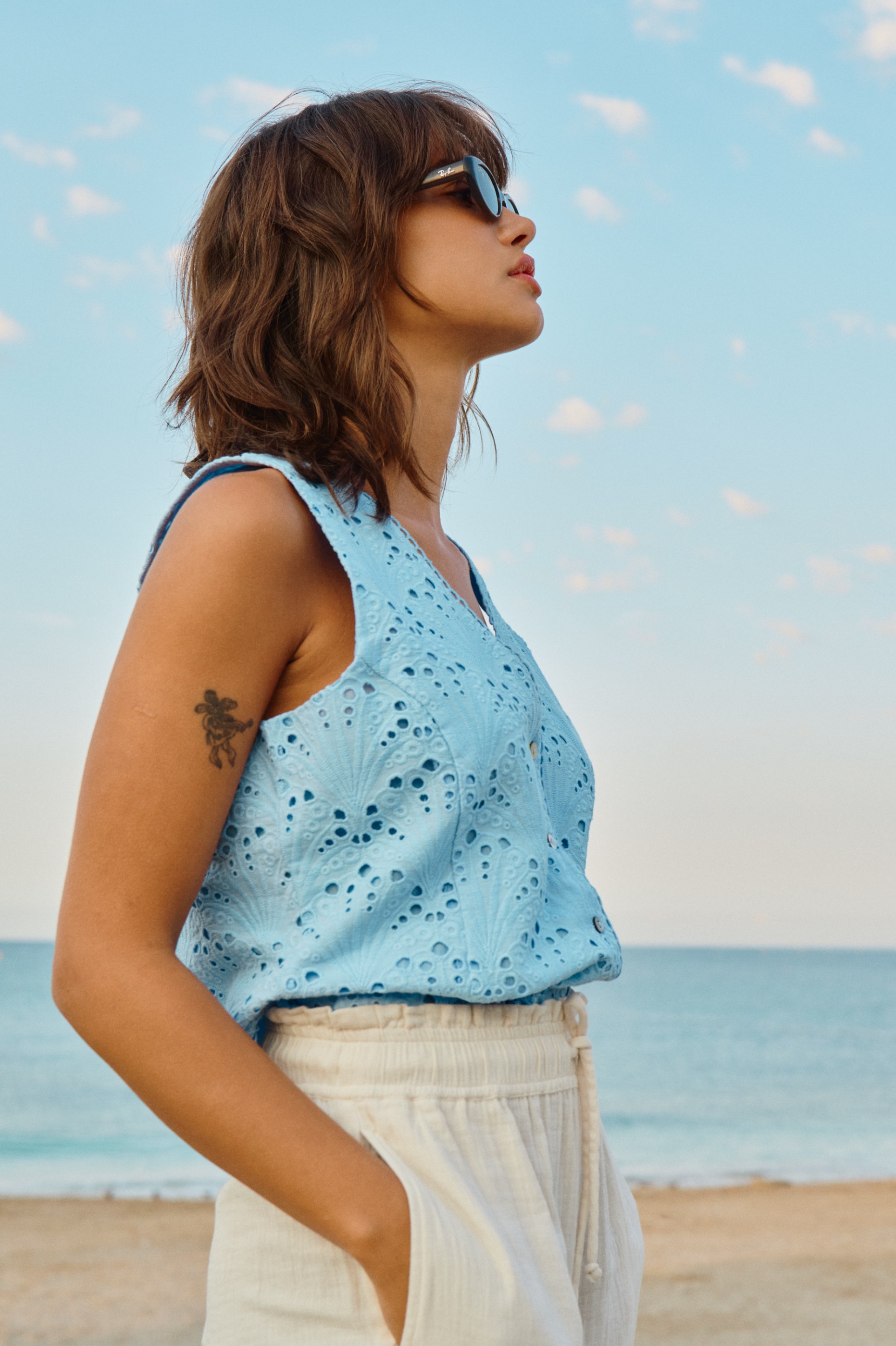 Woman in blue eyelet top, light pants, and Ray-Ban sunglasses on a beach.