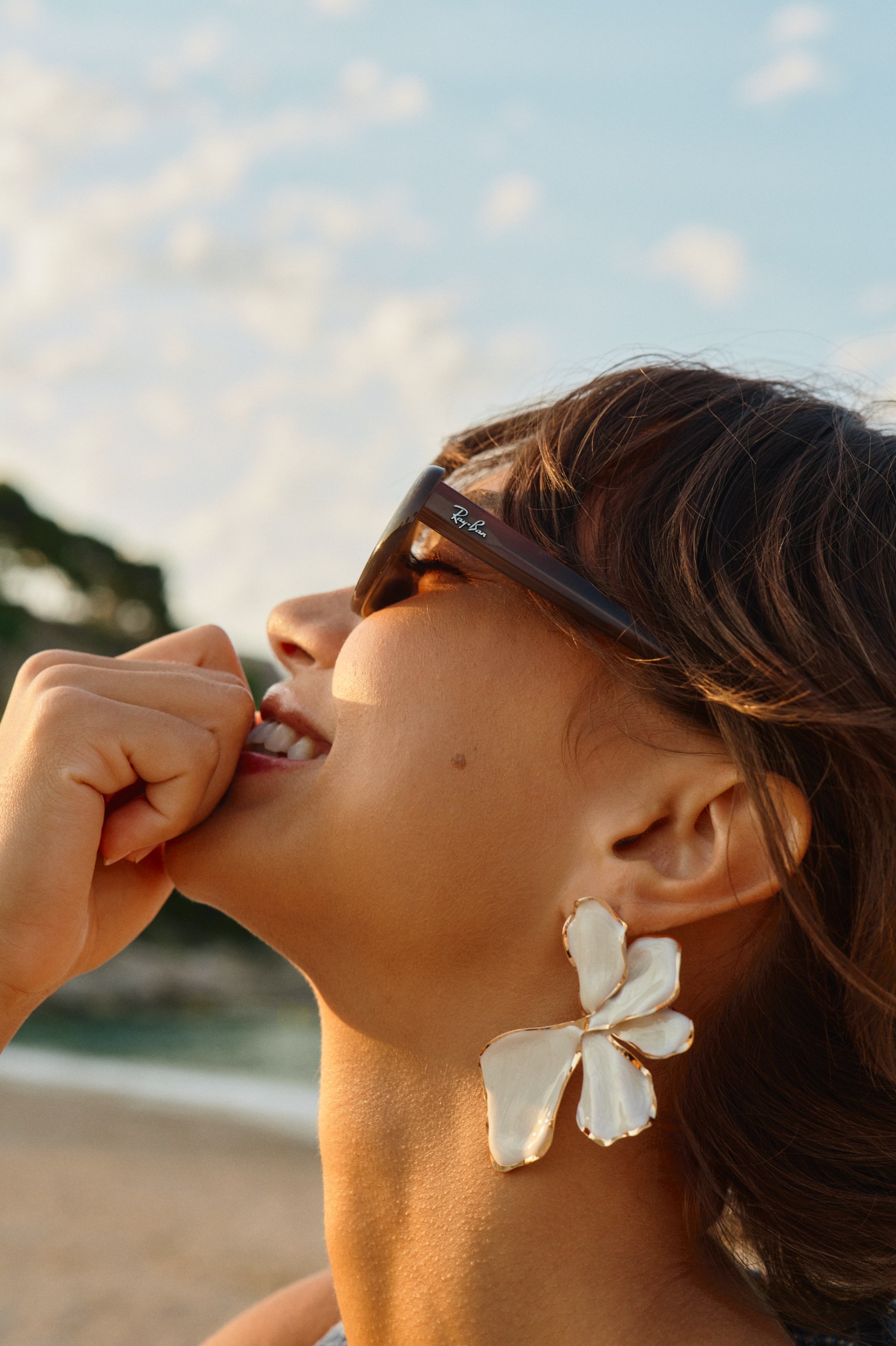 Close-up of a smiling woman in Ray-Ban sunglasses and a large white flower earring.