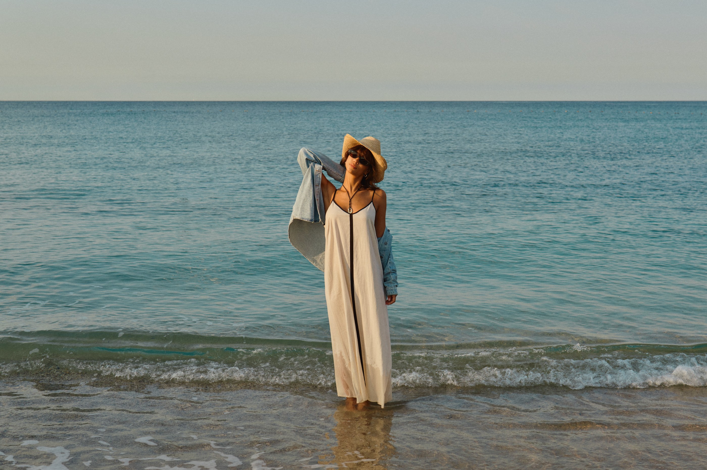 Woman in white dress, denim jacket, straw hat, and sunglasses standing in the ocean.