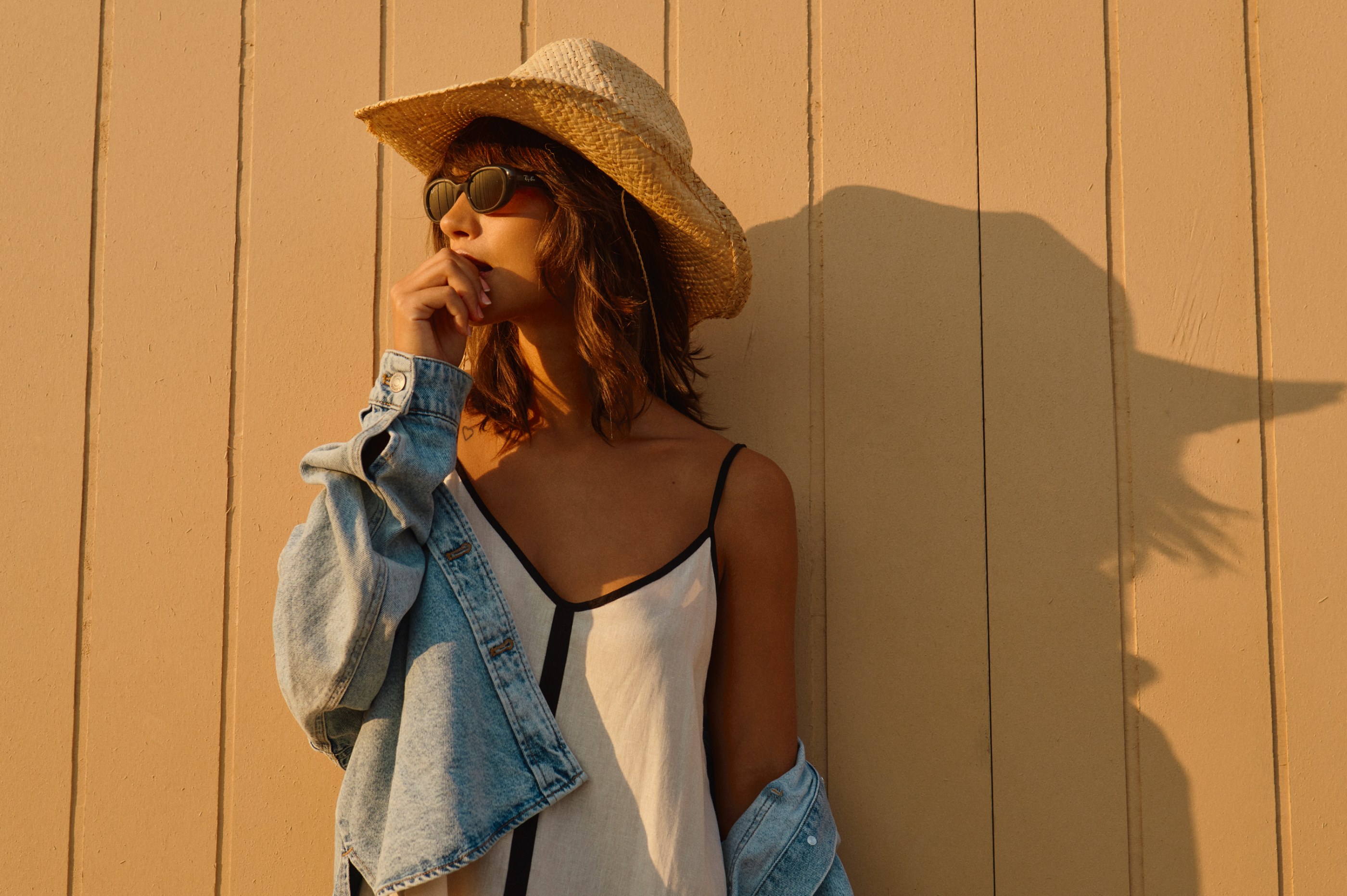 Woman in straw hat, sunglasses, and denim jacket, hand to mouth, against a warm wall.